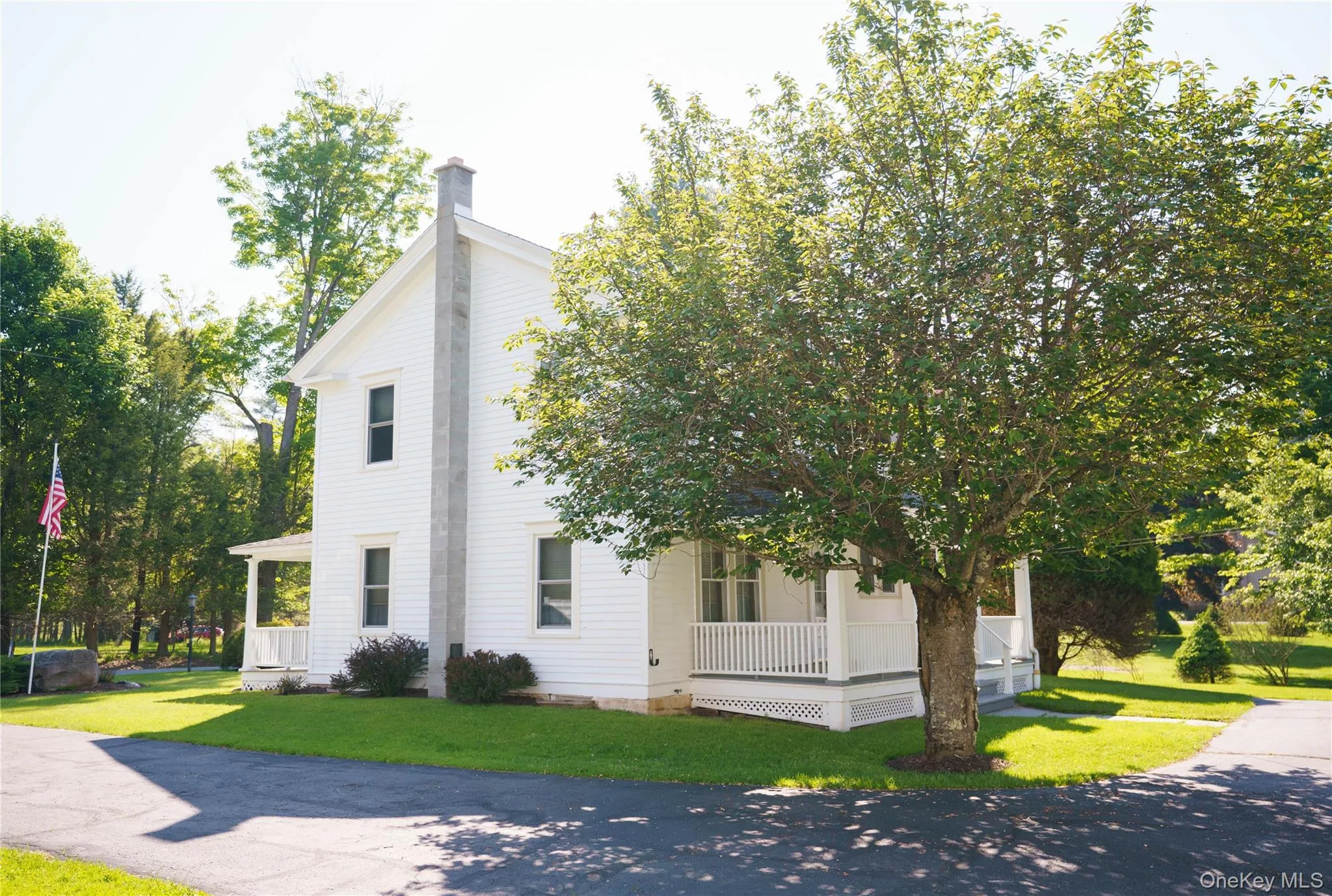White siding exterior featuring a prominent chimney, an inviting front porch with white railings, a paved driveway, and mature landscaping White siding exterior featuring a prominent chimney, an inviting front porch with white railings, a paved driveway, and mature landscaping