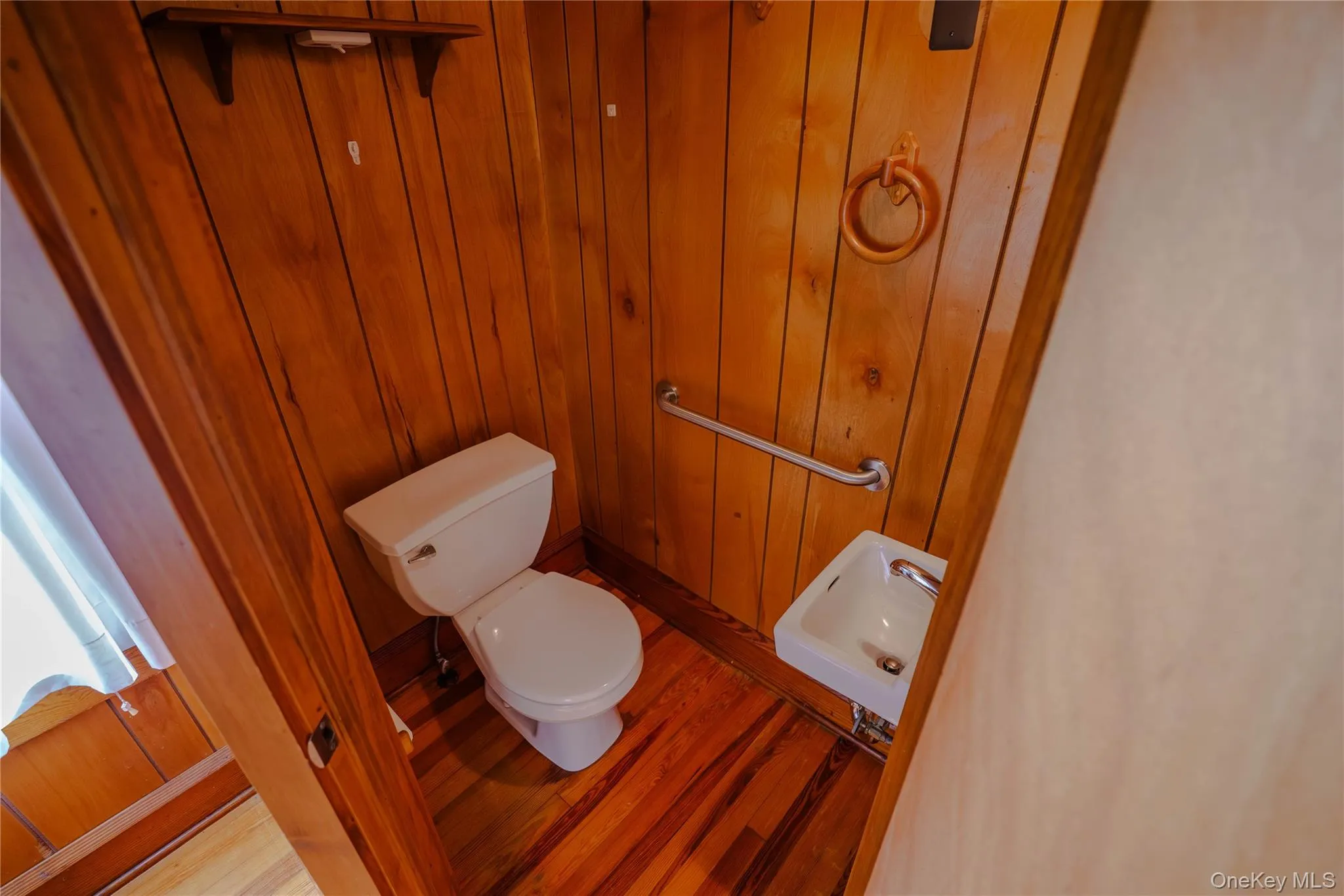 Compact powder room featuring wood-finish paneling, wood-finish flooring, a white toilet, a wall-mounted white sink, and a chrome grab bar Compact powder room featuring wood-finish paneling, wood-finish flooring, a white toilet, a wall-mounted white sink, and a chrome grab bar
