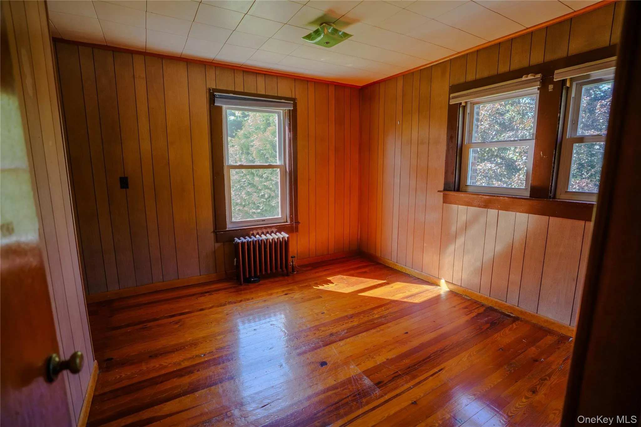 Room featuring wood-finish flooring, vertical wood paneling, two windows with wood trim, and a cast iron radiator Room featuring wood-finish flooring, vertical wood paneling, two windows with wood trim, and a cast iron radiator