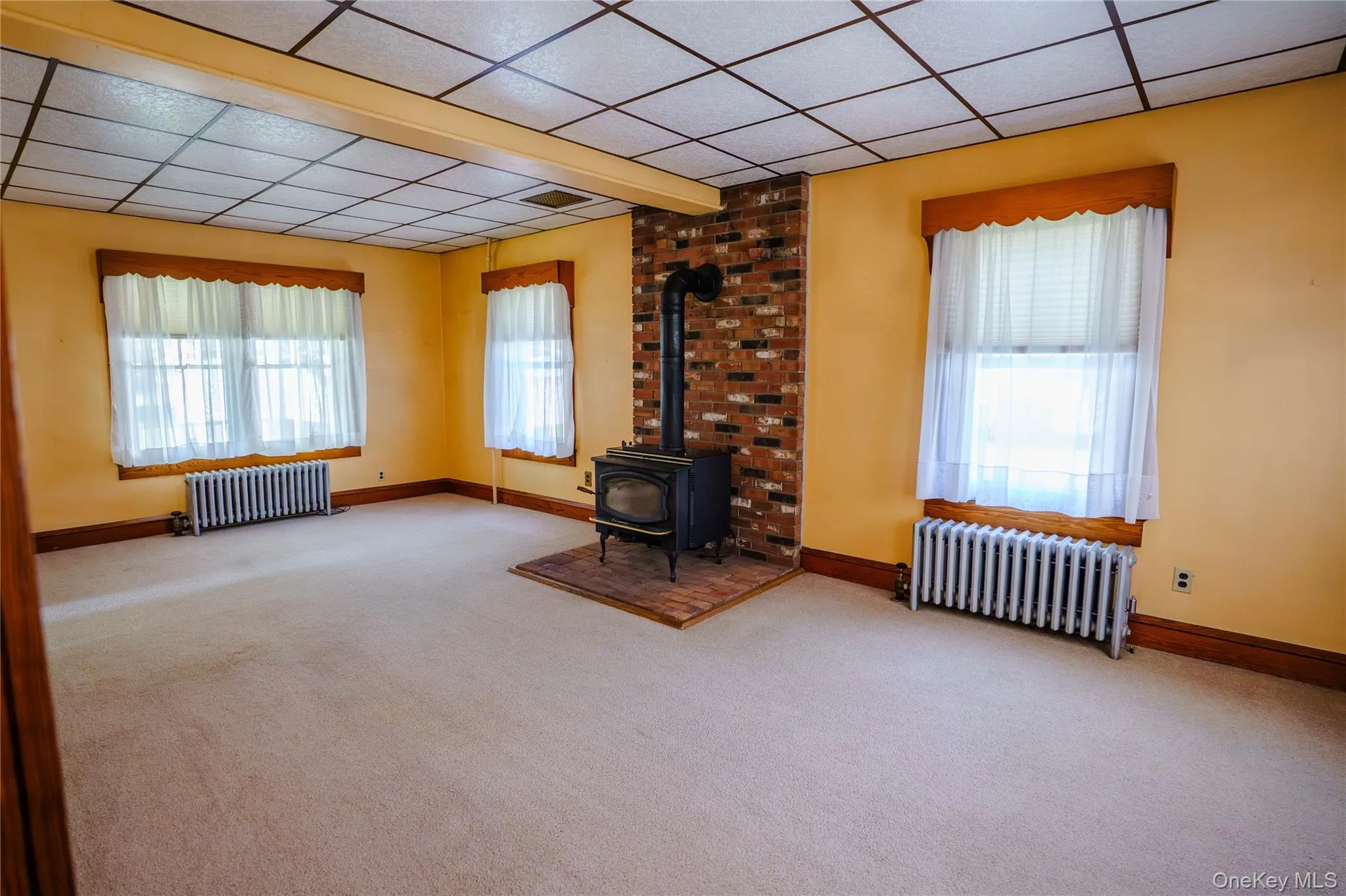 Spacious room featuring a wood-burning stove with a brick surround and hearth, neutral carpet flooring, and two windows with wood trim Spacious room featuring a wood-burning stove with a brick surround and hearth, neutral carpet flooring, and two windows with wood trim