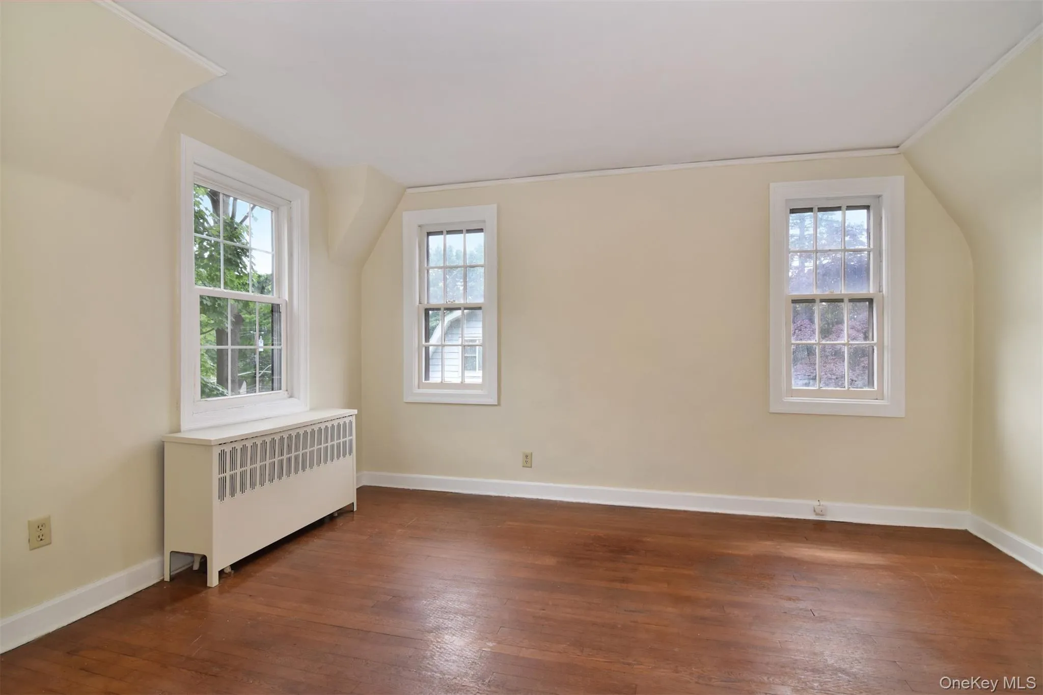 Room featuring three double-hung windows, rich hardwood flooring, and a white radiator cover Room featuring three double-hung windows, rich hardwood flooring, and a white radiator cover