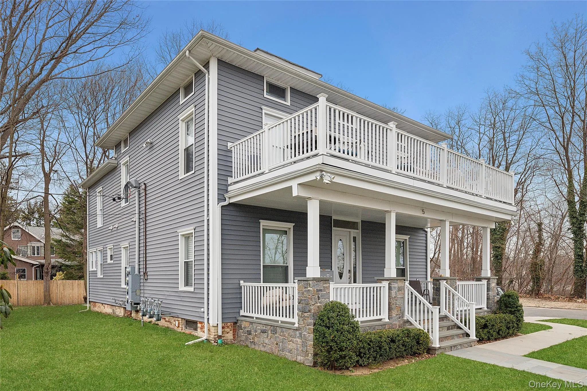 Contemporary residence featuring gray horizontal siding, a stone facade, a covered front porch with white railings, a second-story balcony, and a manicured lawn Contemporary residence featuring gray horizontal siding, a stone facade, a covered front porch with white railings, a second-story balcony, and a manicured lawn