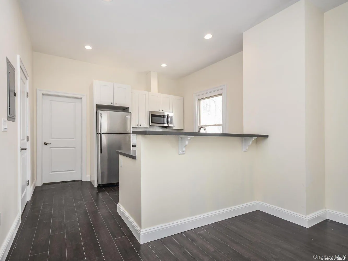 Compact kitchen featuring white cabinetry, stainless steel appliances, dark wood-finish flooring, recessed lighting, and a breakfast bar with a dark countertop Compact kitchen featuring white cabinetry, stainless steel appliances, dark wood-finish flooring, recessed lighting, and a breakfast bar with a dark countertop