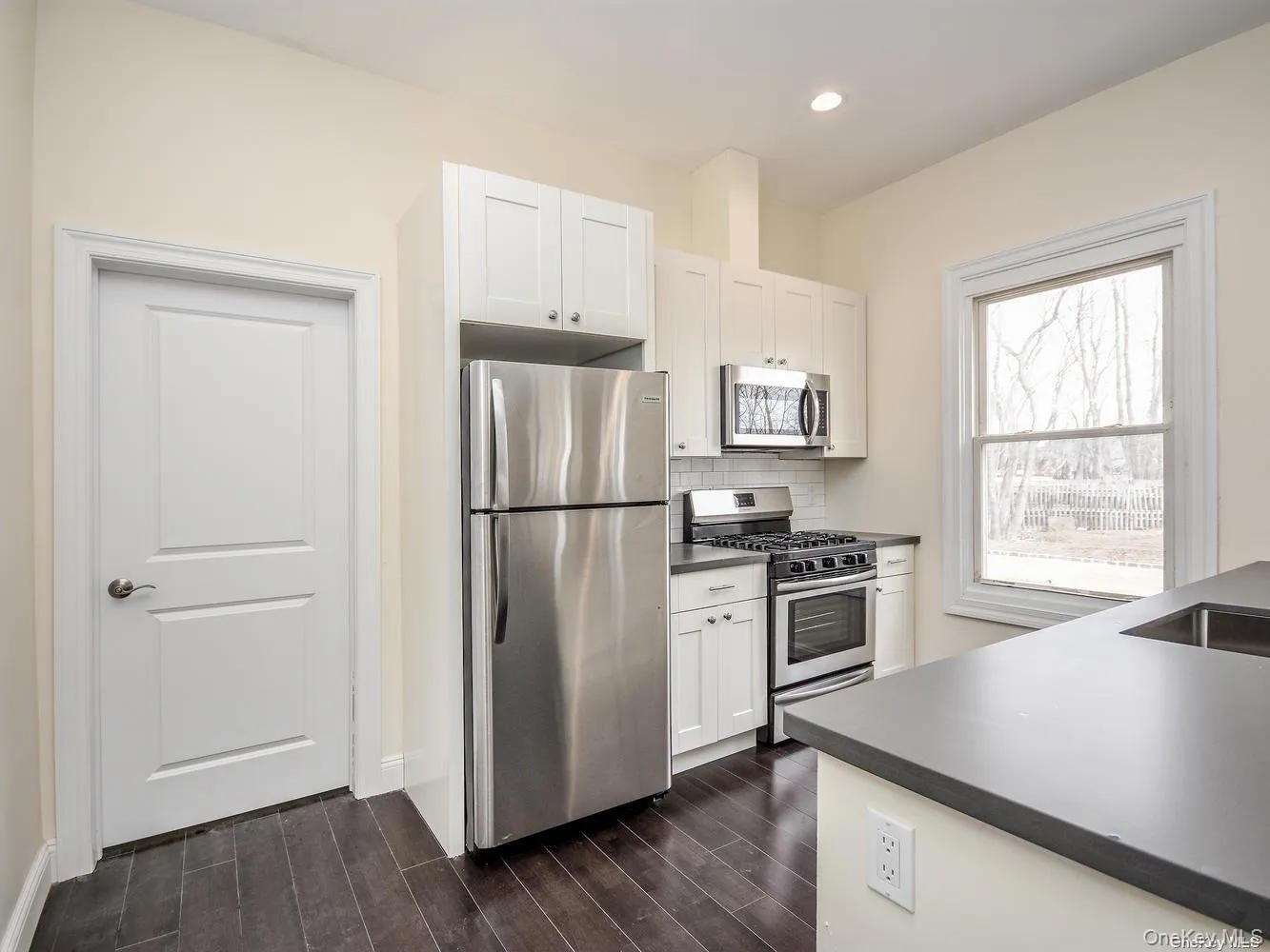 Kitchen featuring white shaker cabinetry, stainless steel appliances, dark wood-finish flooring, and light countertops Kitchen featuring white shaker cabinetry, stainless steel appliances, dark wood-finish flooring, and light countertops