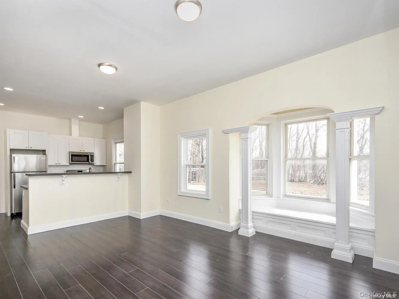 Spacious open-concept room featuring dark wood-finish flooring, a kitchen area with white cabinetry and stainless steel appliances, and a prominent bay window with an integrated window seat Spacious open-concept room featuring dark wood-finish flooring, a kitchen area with white cabinetry and stainless steel appliances, and a prominent bay window with an integrated window seat