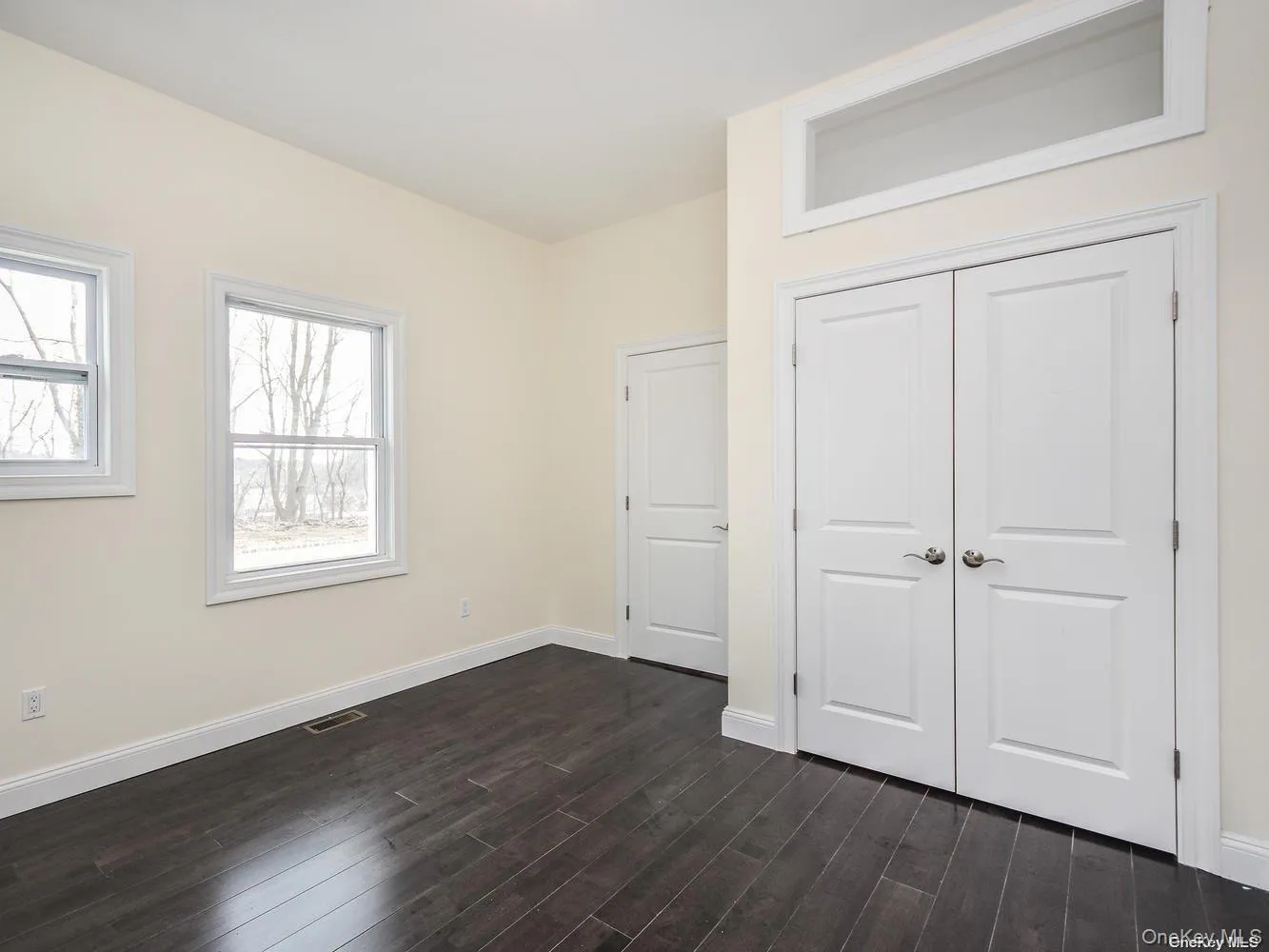 Spacious room with dark wood-finish flooring, two large windows, and a pair of panel doors Spacious room with dark wood-finish flooring, two large windows, and a pair of panel doors