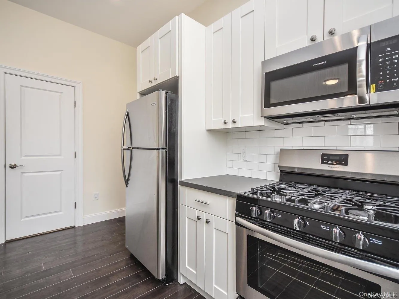 Kitchen featuring stainless steel appliances, white shaker cabinetry, white subway tile backsplash, dark countertops, and wood-finish flooring Kitchen featuring stainless steel appliances, white shaker cabinetry, white subway tile backsplash, dark countertops, and wood-finish flooring