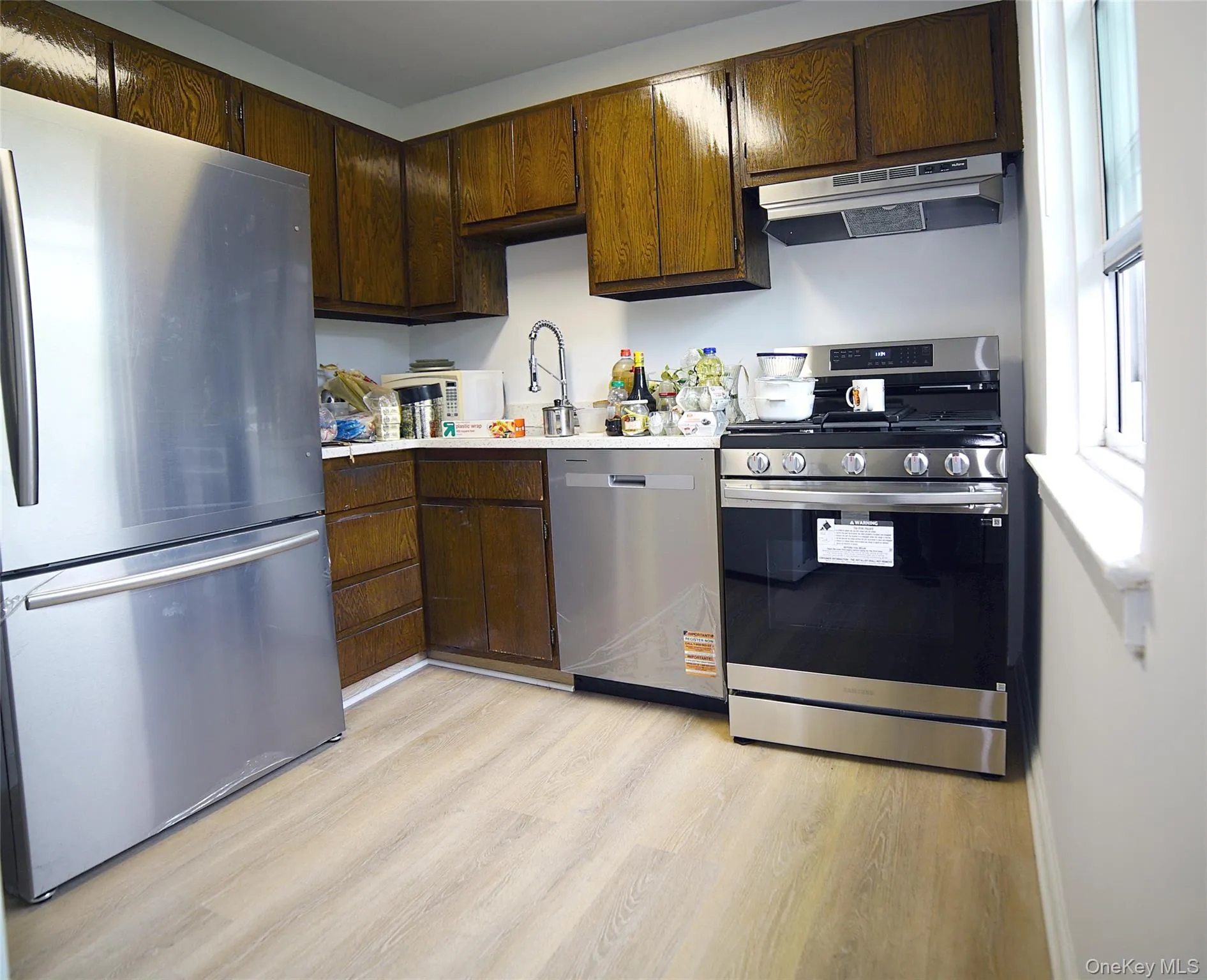 Kitchen featuring light wood-style flooring, stainless steel appliances, under cabinet range hood, a sink, and light countertops Kitchen featuring light wood-style flooring, stainless steel appliances, under cabinet range hood, a sink, and light countertops
