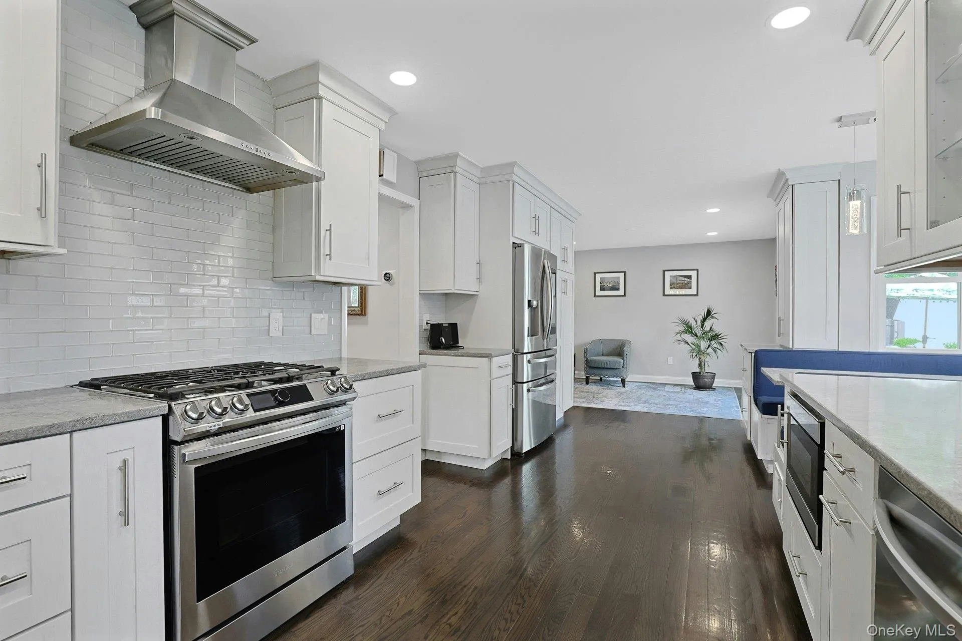 Modern kitchen featuring stainless steel appliances, white shaker cabinetry, an expansive island with built-in seating, light-colored countertops, and dark wood-finish flooring Modern kitchen featuring stainless steel appliances, white shaker cabinetry, an expansive island with built-in seating, light-colored countertops, and dark wood-finish flooring