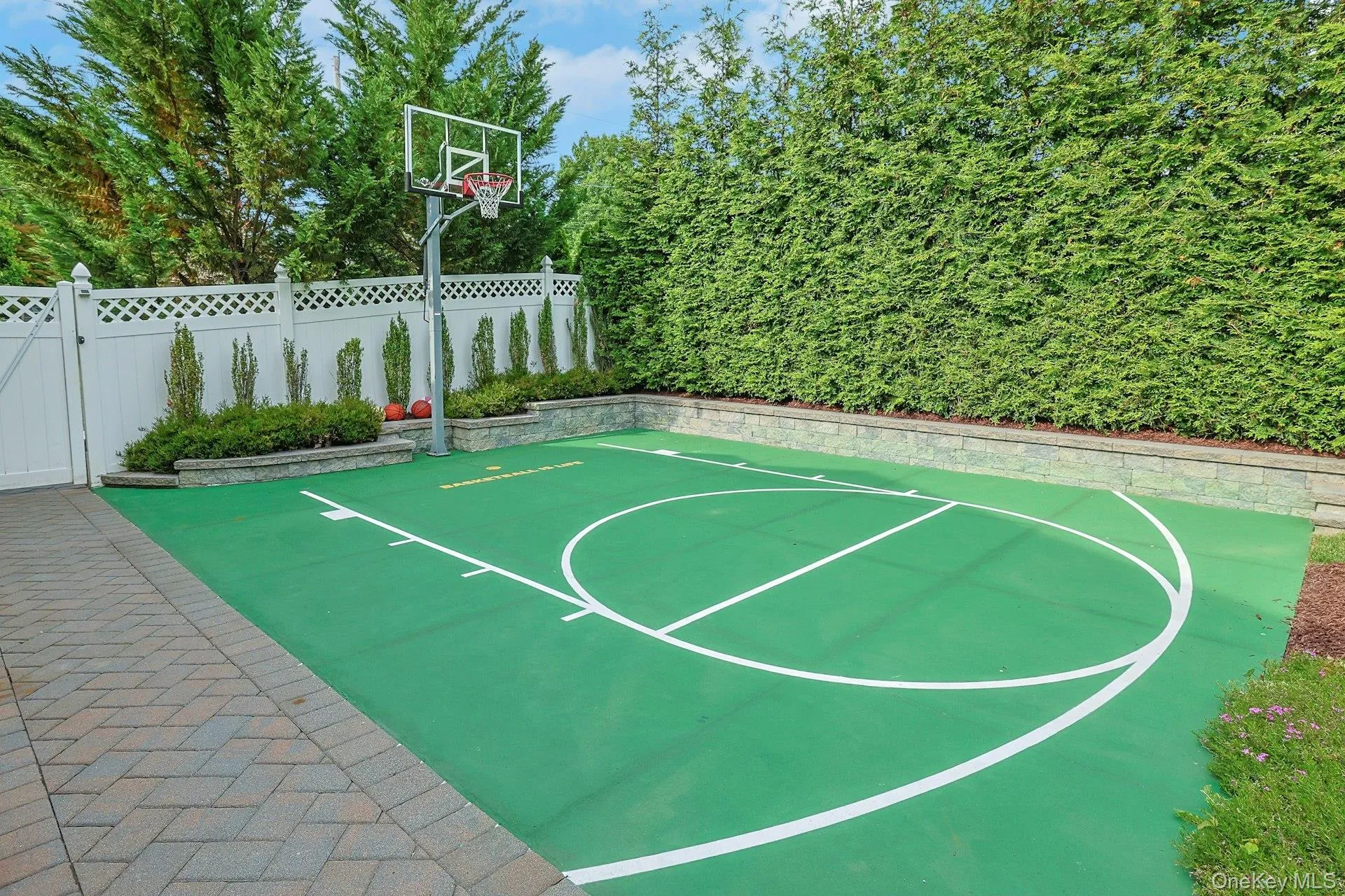 Dedicated sports court featuring a regulation-height basketball hoop, green surface with white court markings, paver stone border, and mature evergreen privacy hedge Dedicated sports court featuring a regulation-height basketball hoop, green surface with white court markings, paver stone border, and mature evergreen privacy hedge