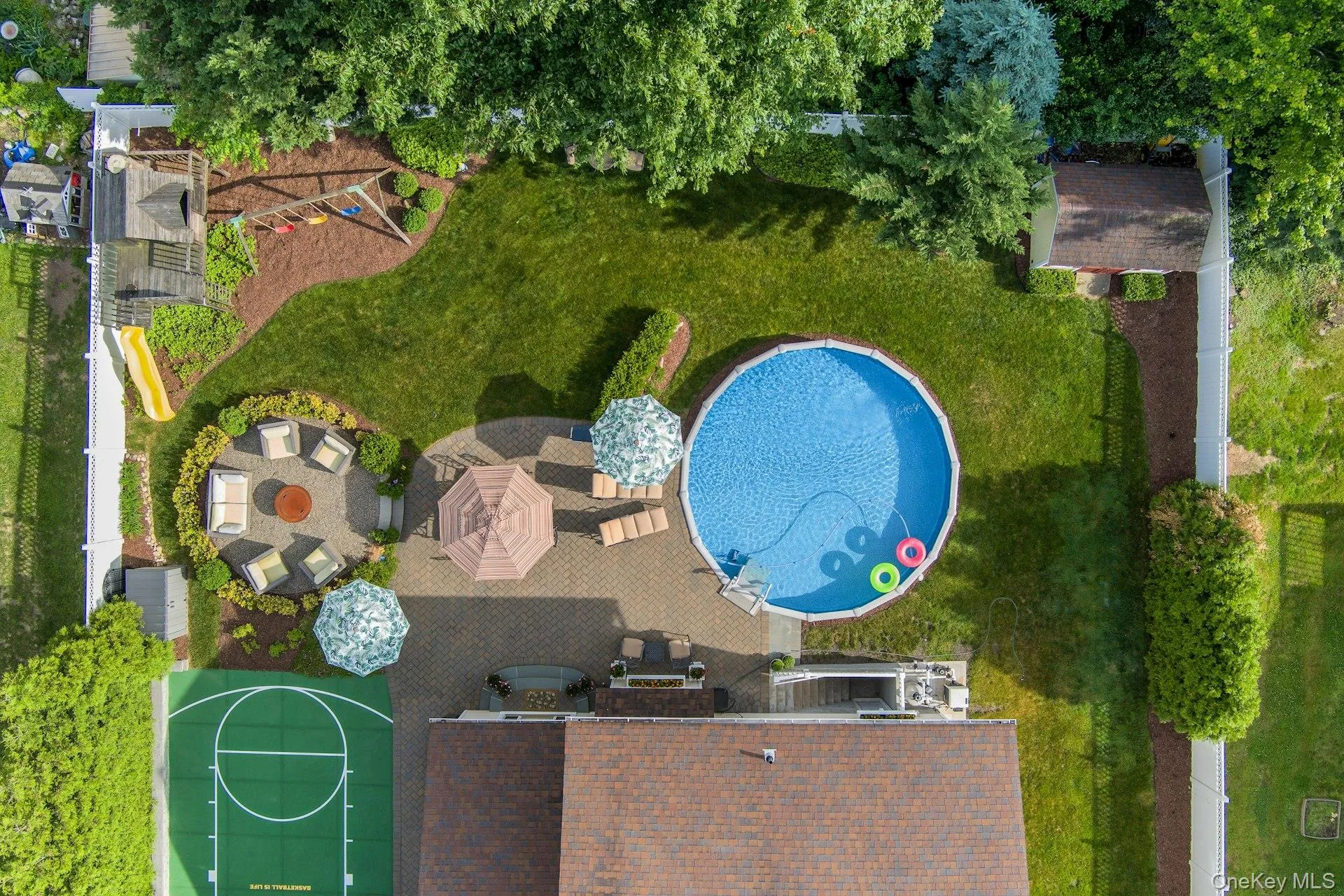 Expansive backyard featuring a large heated swimming pool, a dedicated basketball court, a stone patio with a fire pit area, a playground structure, and lush green lawn Expansive backyard featuring a large heated swimming pool, a dedicated basketball court, a stone patio with a fire pit area, a playground structure, and lush green lawn