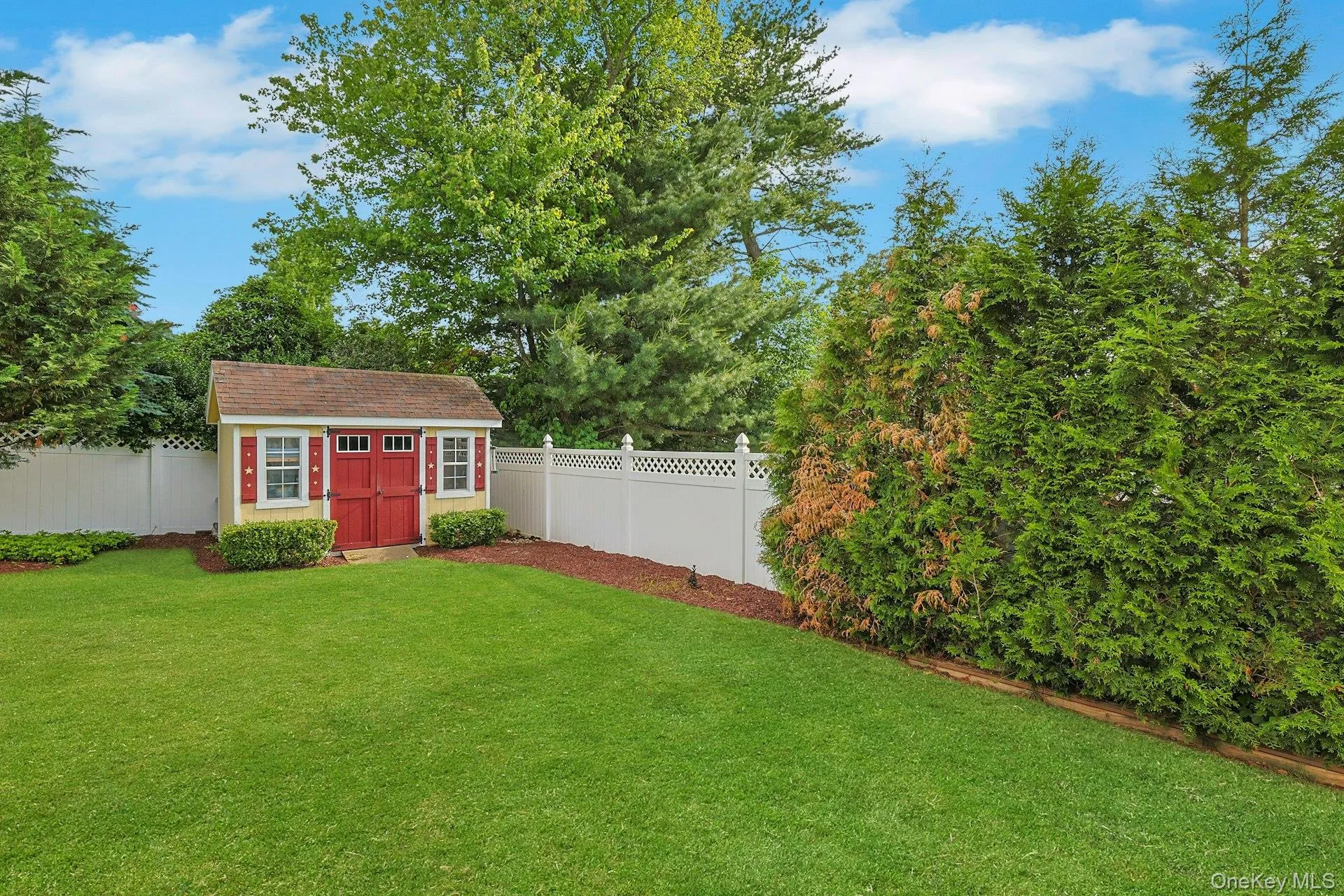 Expansive backyard featuring a manicured lawn, a charming storage shed with red double doors, a white lattice-top fence, and mature perimeter landscaping Expansive backyard featuring a manicured lawn, a charming storage shed with red double doors, a white lattice-top fence, and mature perimeter landscaping