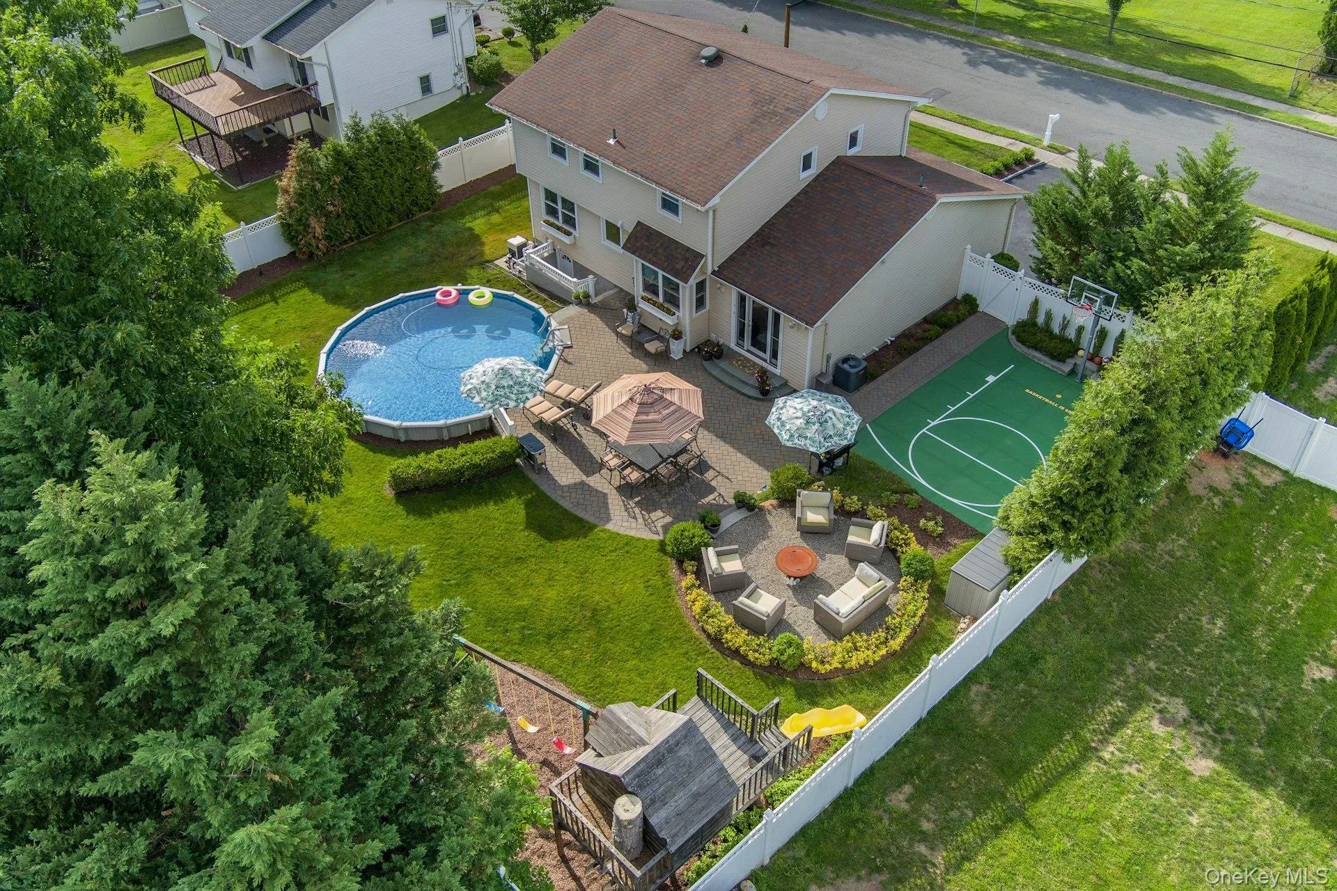 Expansive backyard featuring an above-ground pool, a paved patio, a dedicated sports court, a circular fire pit area, and mature perimeter trees Expansive backyard featuring an above-ground pool, a paved patio, a dedicated sports court, a circular fire pit area, and mature perimeter trees