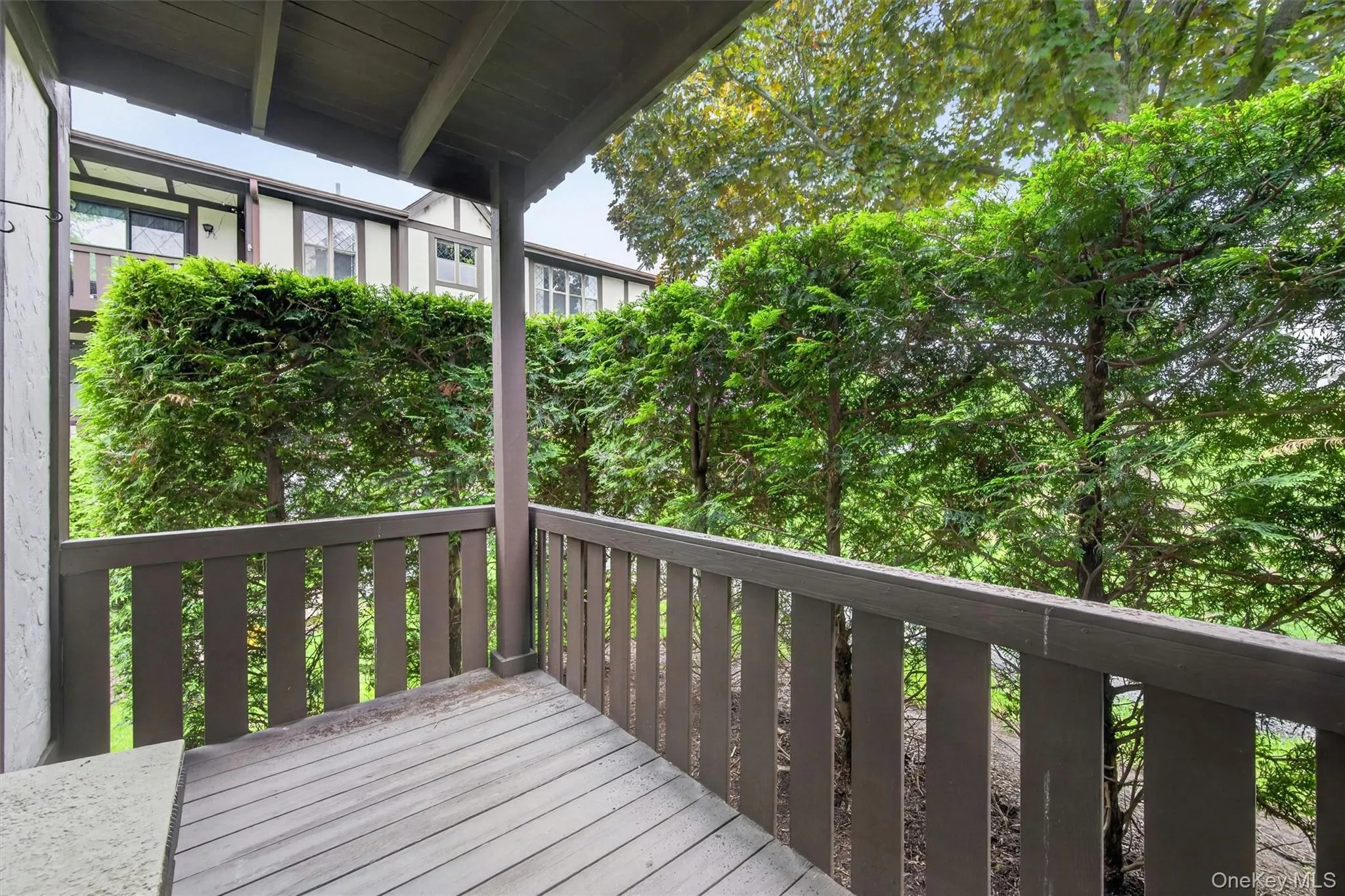 Covered porch with wood-finish decking and railing, surrounded by mature trees and manicured hedges Covered porch with wood-finish decking and railing, surrounded by mature trees and manicured hedges