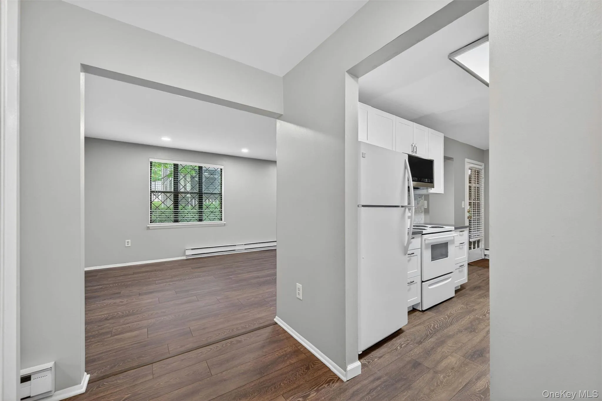Open-concept living area featuring wood-finish flooring, a large window with decorative grilles, and baseboard heating Open-concept living area featuring wood-finish flooring, a large window with decorative grilles, and baseboard heating
