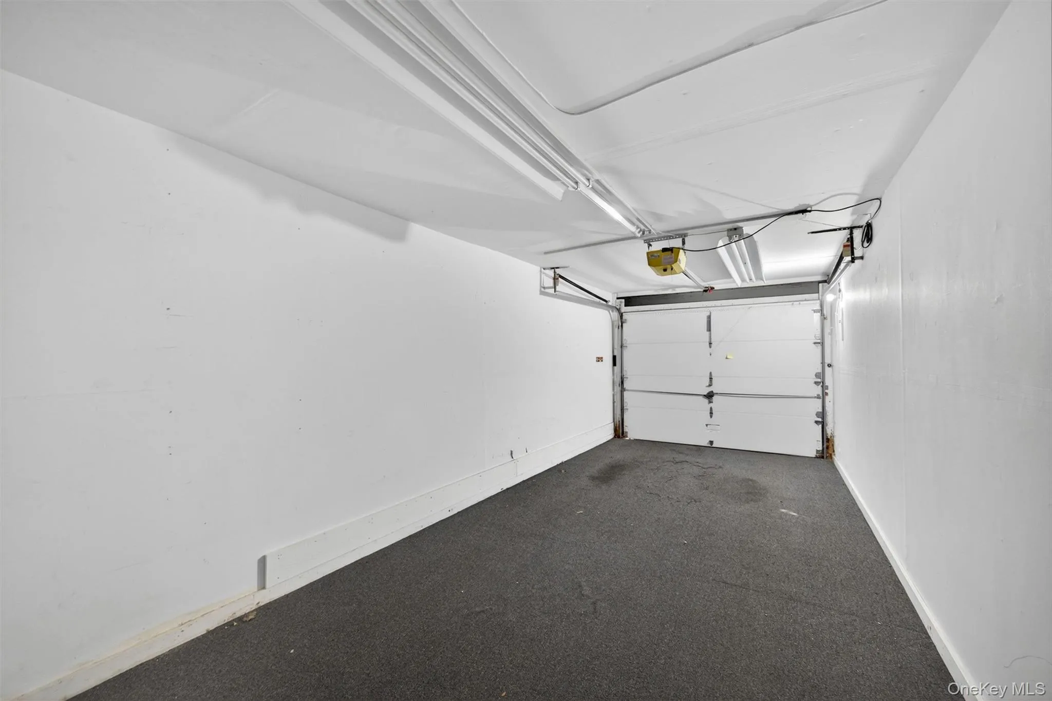 Single-car garage featuring a white sectional garage door, painted white walls, track lighting, and a dark textured floor Single-car garage featuring a white sectional garage door, painted white walls, track lighting, and a dark textured floor