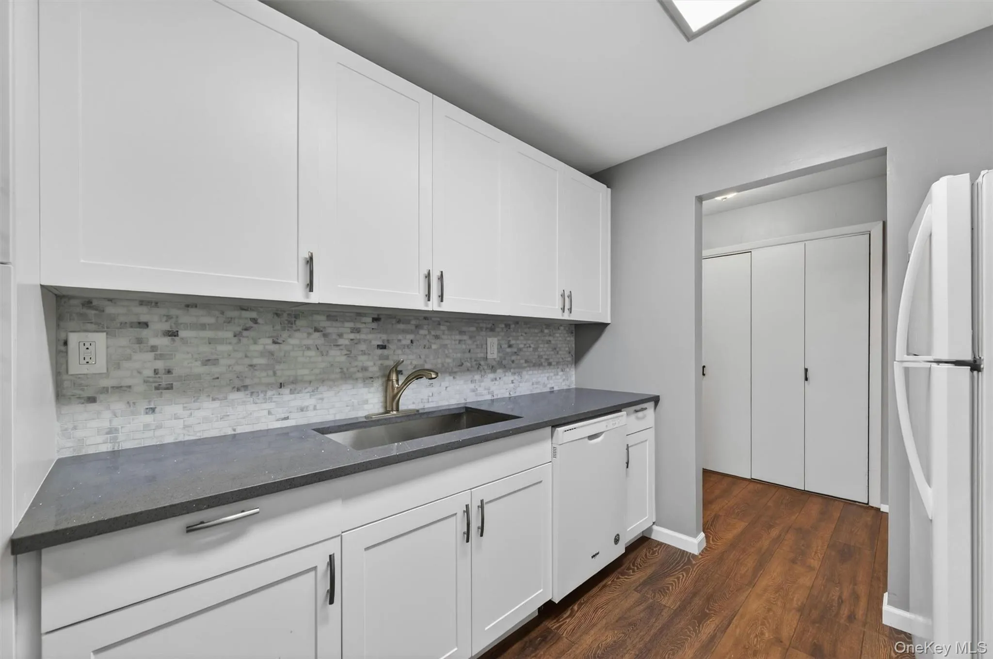 Kitchen featuring white cabinetry with brushed nickel hardware, a stainless steel sink, a mosaic tile backsplash, dark grey countertops, and wood-finish flooring Kitchen featuring white cabinetry with brushed nickel hardware, a stainless steel sink, a mosaic tile backsplash, dark grey countertops, and wood-finish flooring