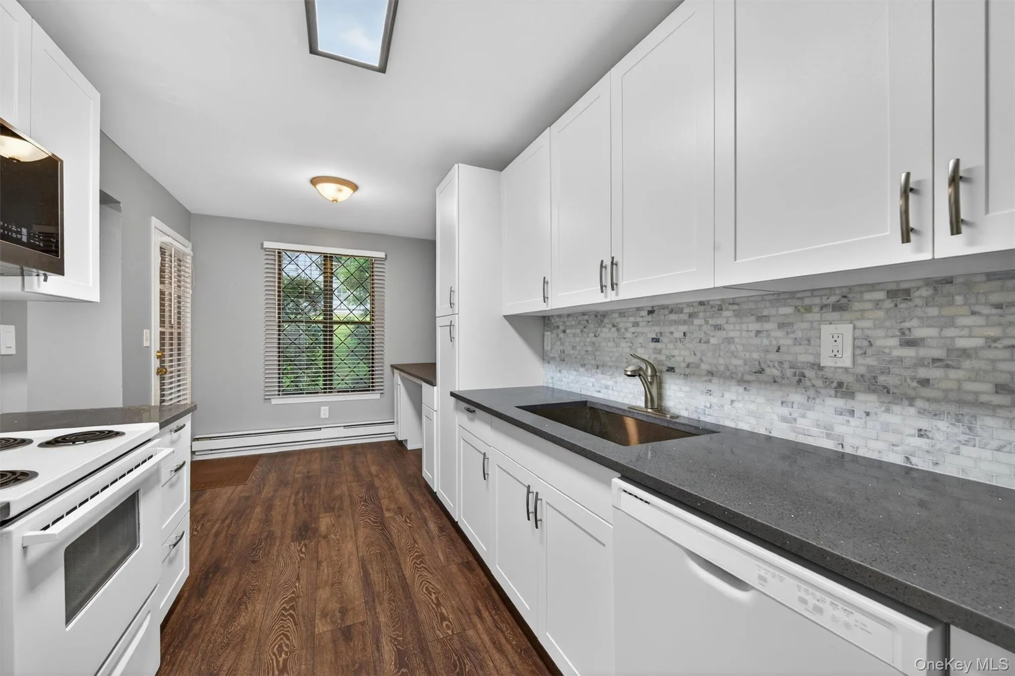 Modern kitchen featuring white shaker-style cabinetry, dark gray countertops, a subway tile backsplash, wood-finish flooring, and a ceiling skylight Modern kitchen featuring white shaker-style cabinetry, dark gray countertops, a subway tile backsplash, wood-finish flooring, and a ceiling skylight