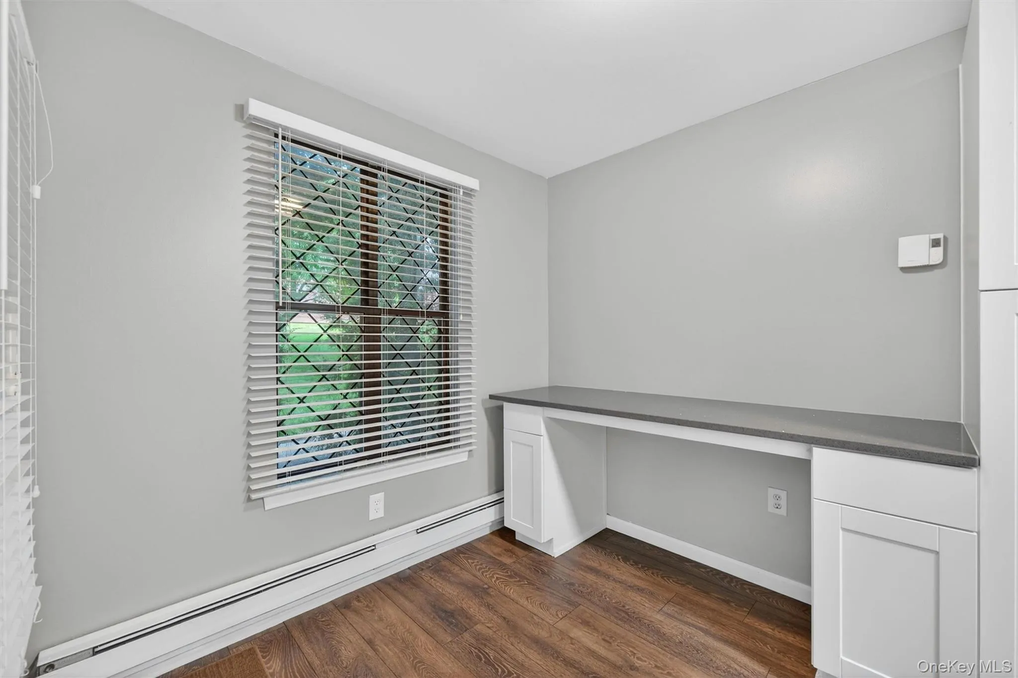 Built-in desk with a dark countertop and white cabinetry, wood-finish flooring, and a window with horizontal blinds and security bars Built-in desk with a dark countertop and white cabinetry, wood-finish flooring, and a window with horizontal blinds and security bars