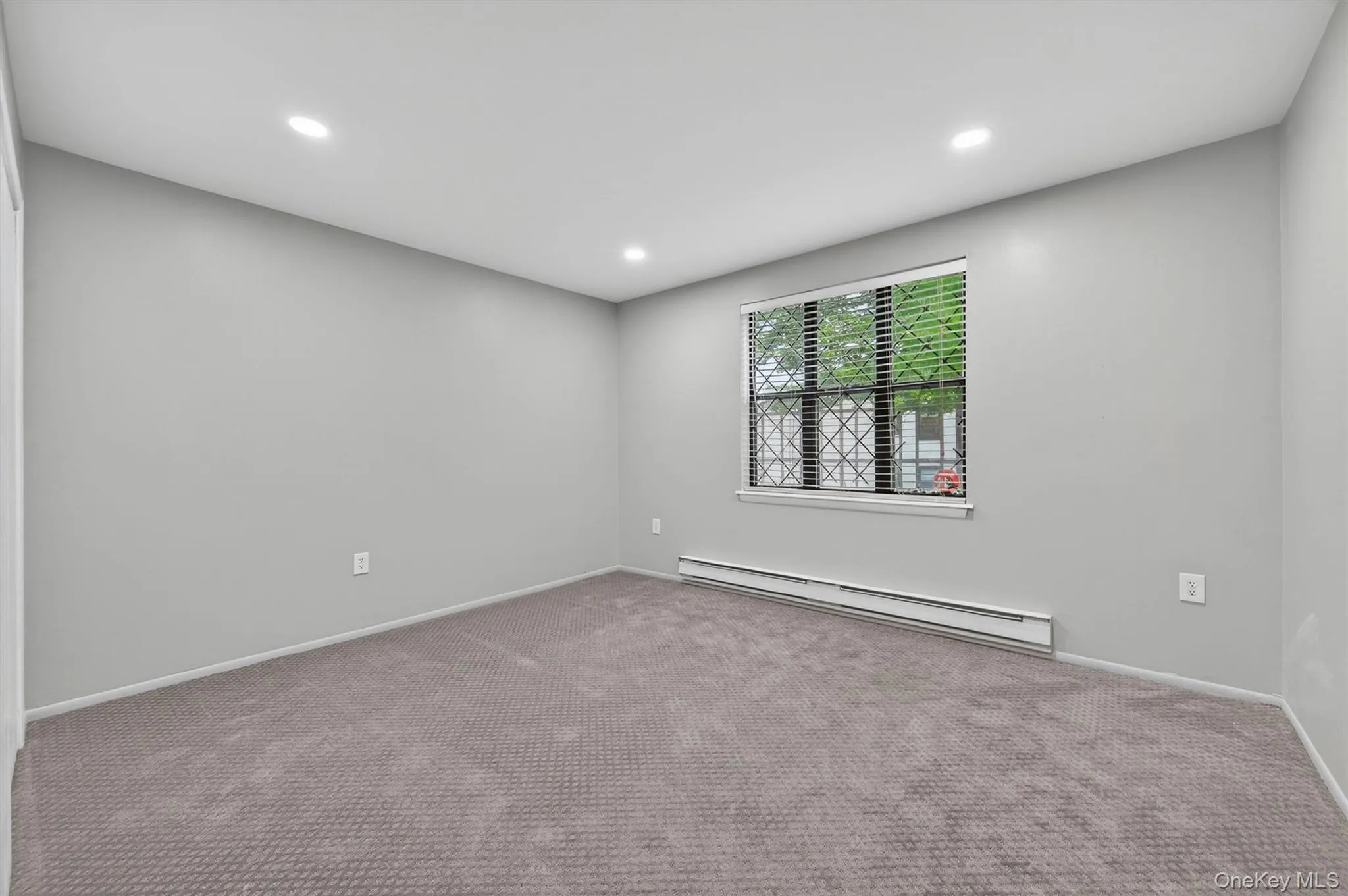 Interior space featuring a window with horizontal blinds and security bars, wall-to-wall carpeting, recessed ceiling lighting, light gray painted walls, and baseboard heating Interior space featuring a window with horizontal blinds and security bars, wall-to-wall carpeting, recessed ceiling lighting, light gray painted walls, and baseboard heating