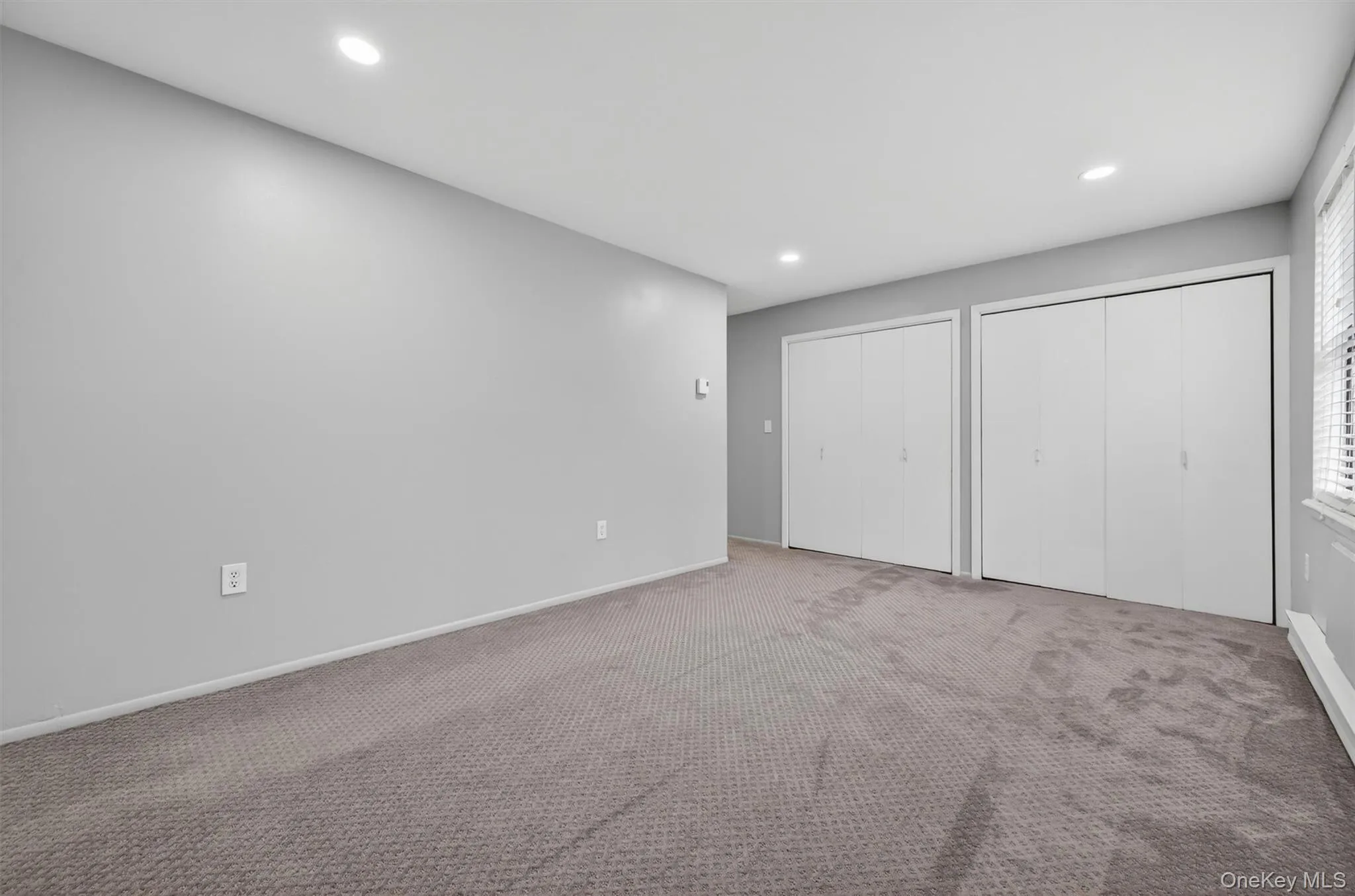 Carpeted room featuring light gray walls, two sets of white bi-fold closet doors, recessed lighting, and a window with white blinds Carpeted room featuring light gray walls, two sets of white bi-fold closet doors, recessed lighting, and a window with white blinds