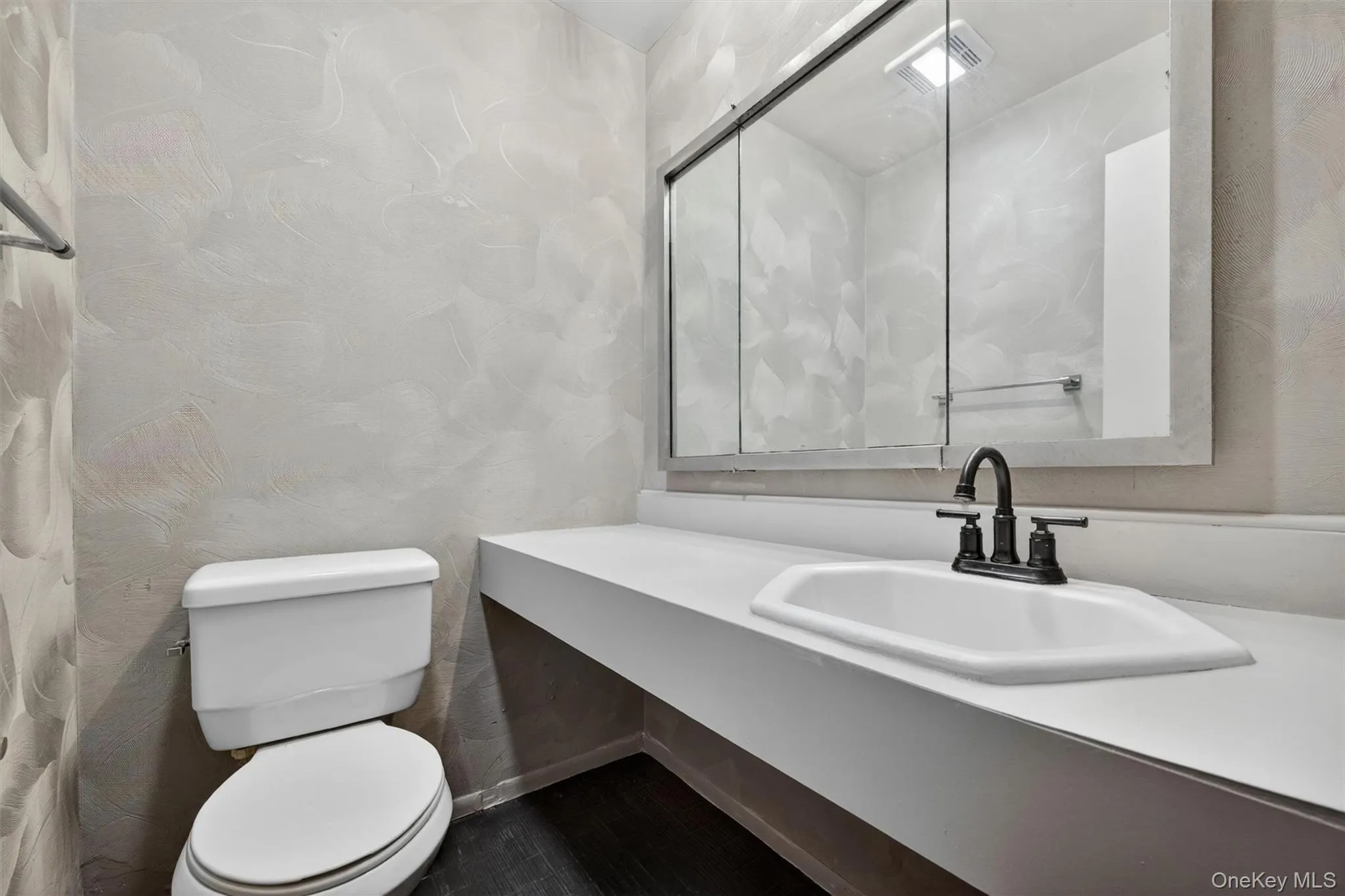 Bathroom featuring a white elongated toilet, dark wood-finish flooring, and light-toned textured walls Bathroom featuring a white elongated toilet, dark wood-finish flooring, and light-toned textured walls