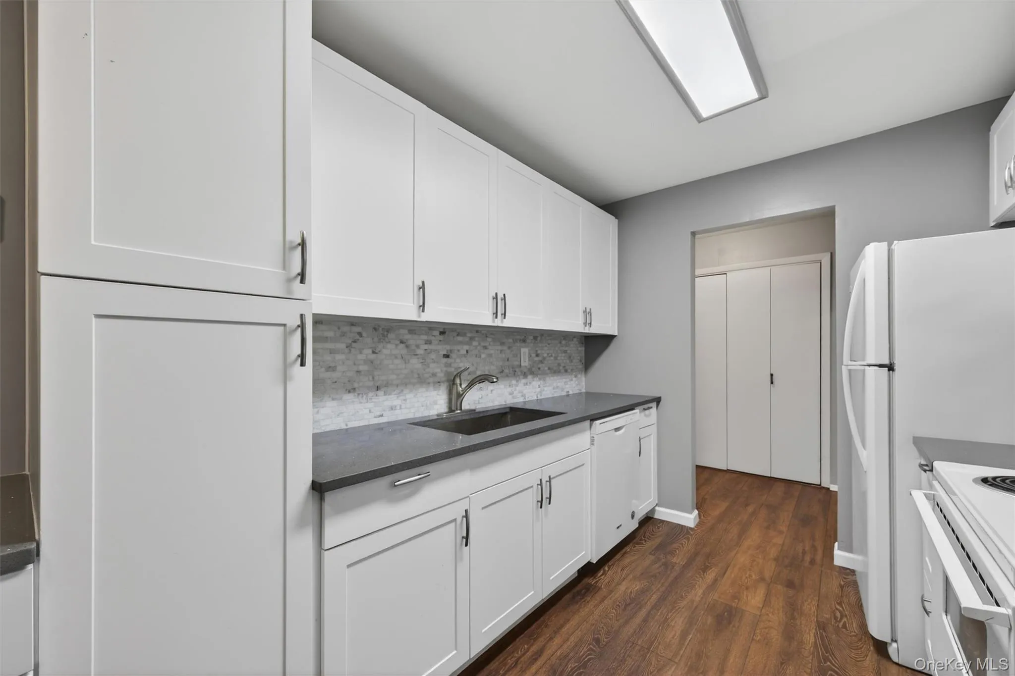 Modern kitchen featuring white shaker cabinetry, dark countertops, a mosaic tile backsplash, and wood-finish flooring Modern kitchen featuring white shaker cabinetry, dark countertops, a mosaic tile backsplash, and wood-finish flooring