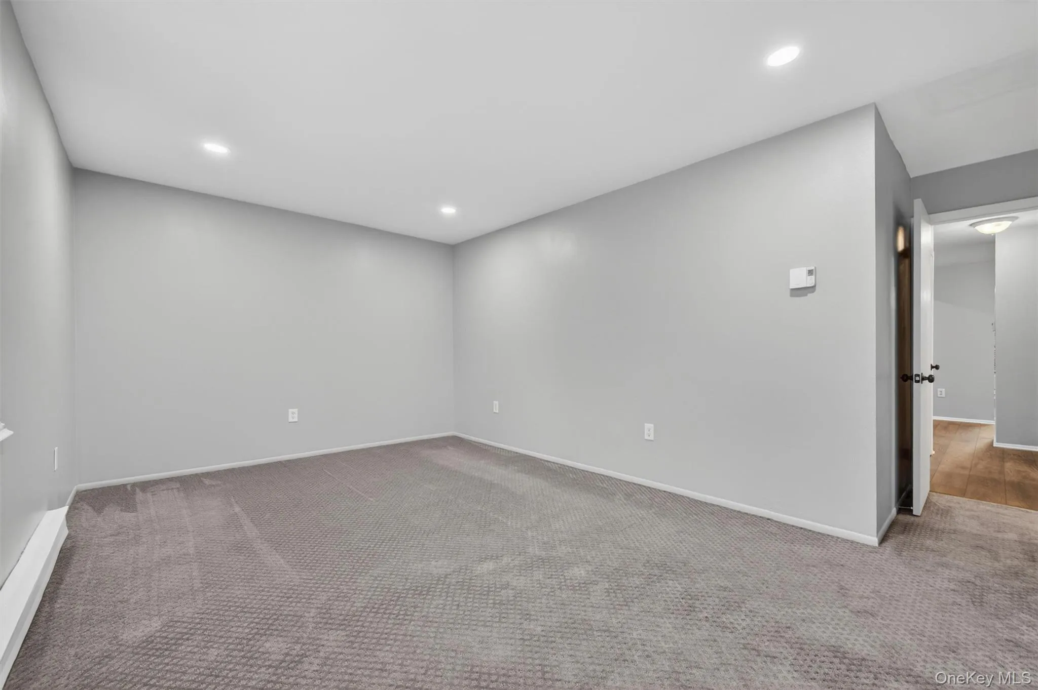 Spacious room featuring neutral gray walls, recessed lighting, gray patterned carpeting, white baseboards, and a doorway leading to a wood-finish flooring area Spacious room featuring neutral gray walls, recessed lighting, gray patterned carpeting, white baseboards, and a doorway leading to a wood-finish flooring area
