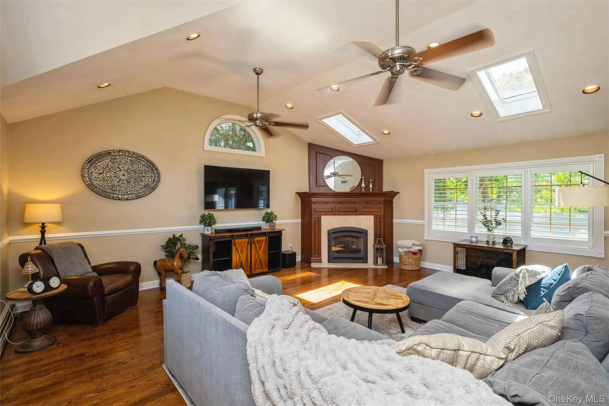 Spacious great room featuring vaulted ceilings with skylights, recessed lighting, wood-finish flooring, a gas fireplace with a mantel, and multiple windows with plantation shutters Spacious great room featuring vaulted ceilings with skylights, recessed lighting, wood-finish flooring, a gas fireplace with a mantel, and multiple windows with plantation shutters