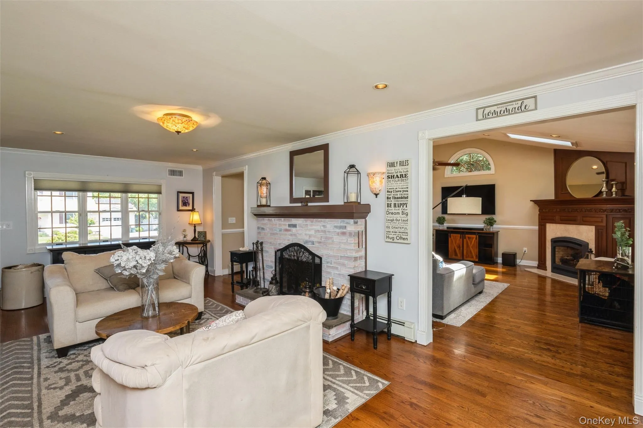 Living area featuring wood-finish flooring, a painted brick fireplace with a wood mantle, and a decorative ceiling light fixture Living area featuring wood-finish flooring, a painted brick fireplace with a wood mantle, and a decorative ceiling light fixture
