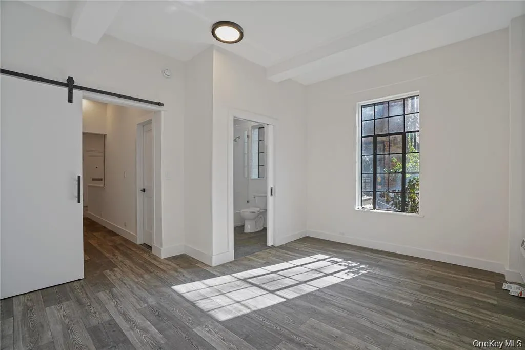 Spacious room featuring wood-finish flooring, a large multi-pane window, and exposed ceiling beams Spacious room featuring wood-finish flooring, a large multi-pane window, and exposed ceiling beams