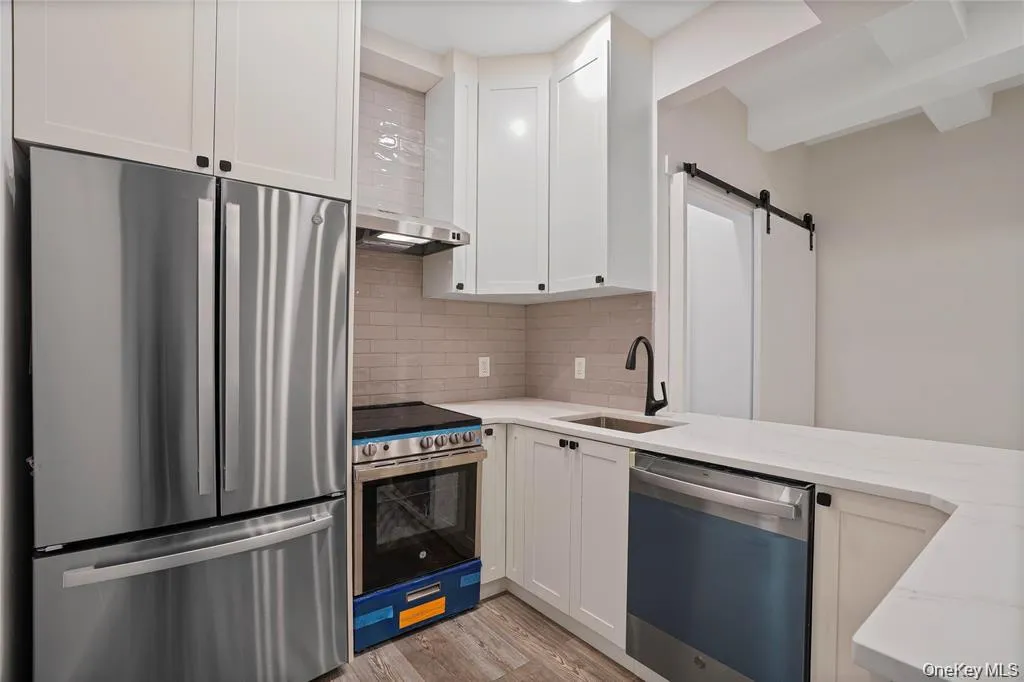 Modern kitchen featuring stainless steel appliances, white shaker cabinetry with black hardware, a light tile backsplash, light countertops, and wood-finish flooring Modern kitchen featuring stainless steel appliances, white shaker cabinetry with black hardware, a light tile backsplash, light countertops, and wood-finish flooring
