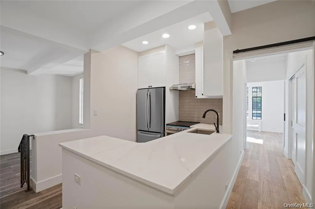 Modern kitchen featuring white cabinetry, a stainless steel refrigerator, a stainless steel range hood, a subway tile backsplash, and a white countertop with an undermount sink and matte black faucet Modern kitchen featuring white cabinetry, a stainless steel refrigerator, a stainless steel range hood, a subway tile backsplash, and a white countertop with an undermount sink and matte black faucet