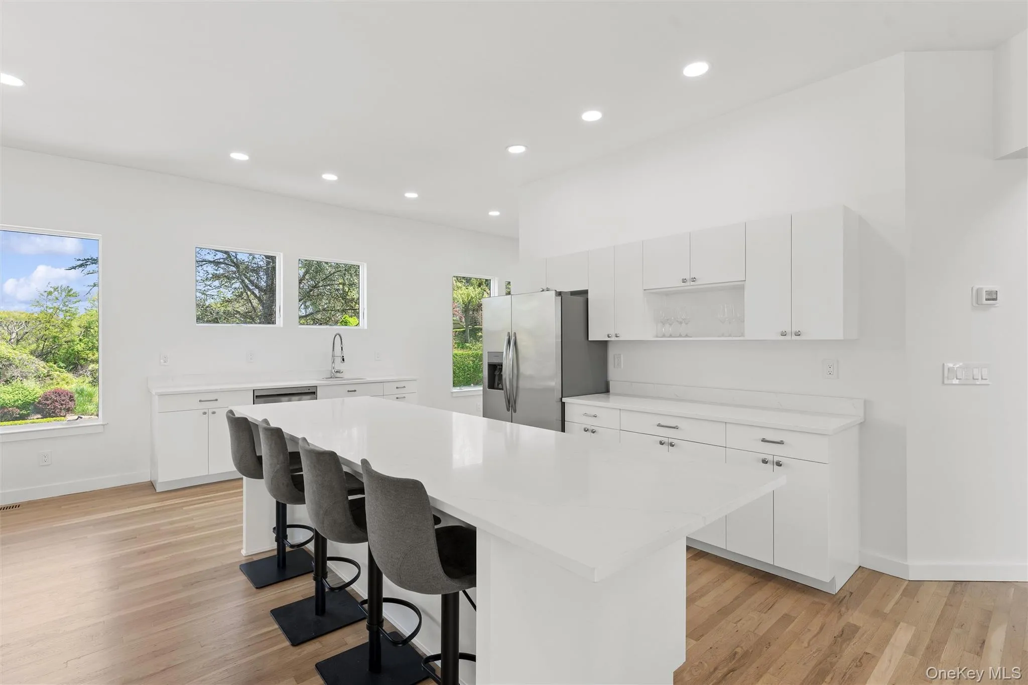 1st Floor Spacious kitchen featuring a large central island with seating, extensive white cabinetry, stainless steel refrigerator, recessed lighting, and wood-finish flooring 1st Floor Spacious kitchen featuring a large central island with seating, extensive white cabinetry, stainless steel refrigerator, recessed lighting, and wood-finish flooring