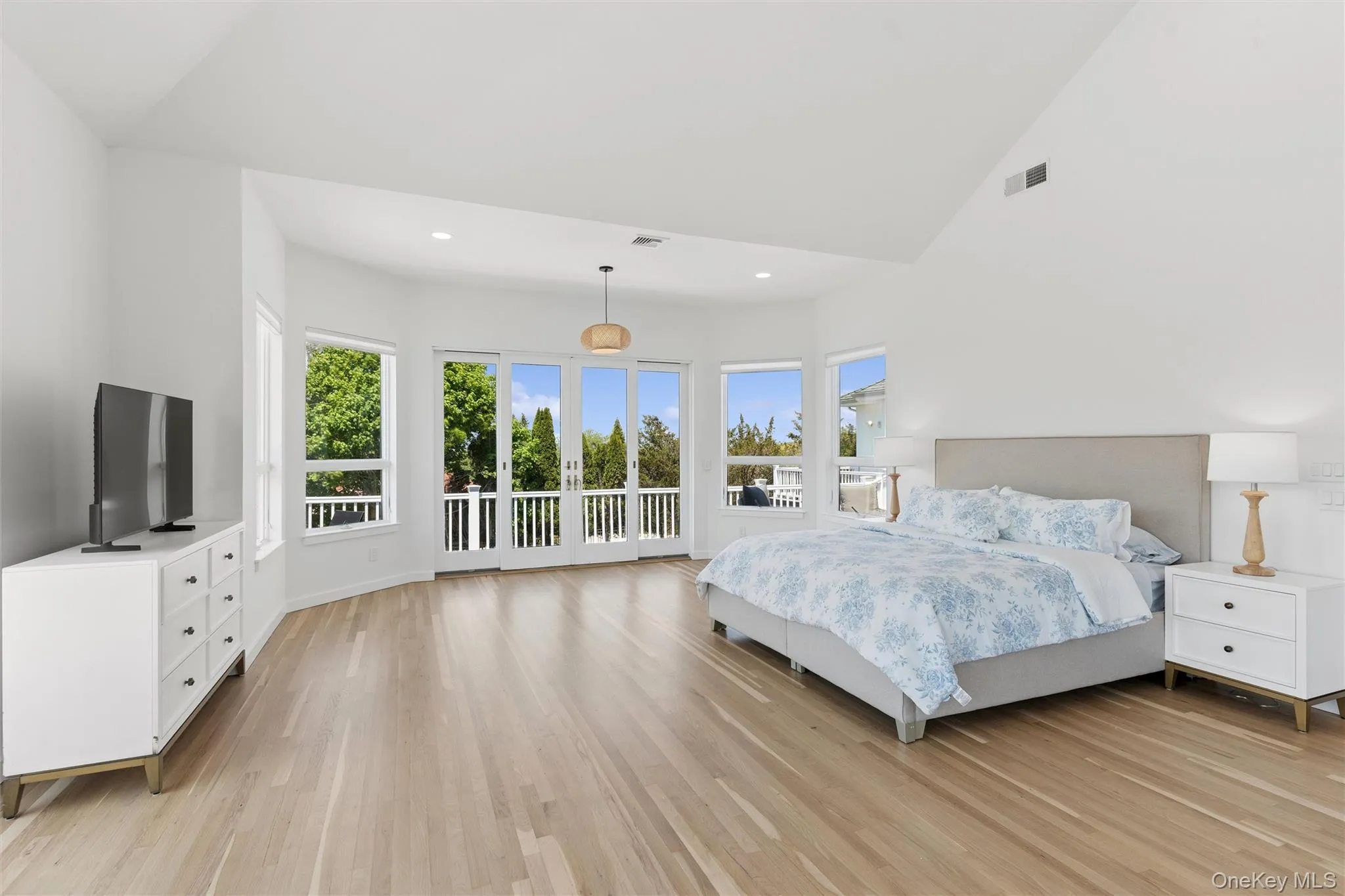 Vaulted ceiling bedroom with light wood-finish flooring, featuring multiple windows and French doors opening to a balcony Vaulted ceiling bedroom with light wood-finish flooring, featuring multiple windows and French doors opening to a balcony