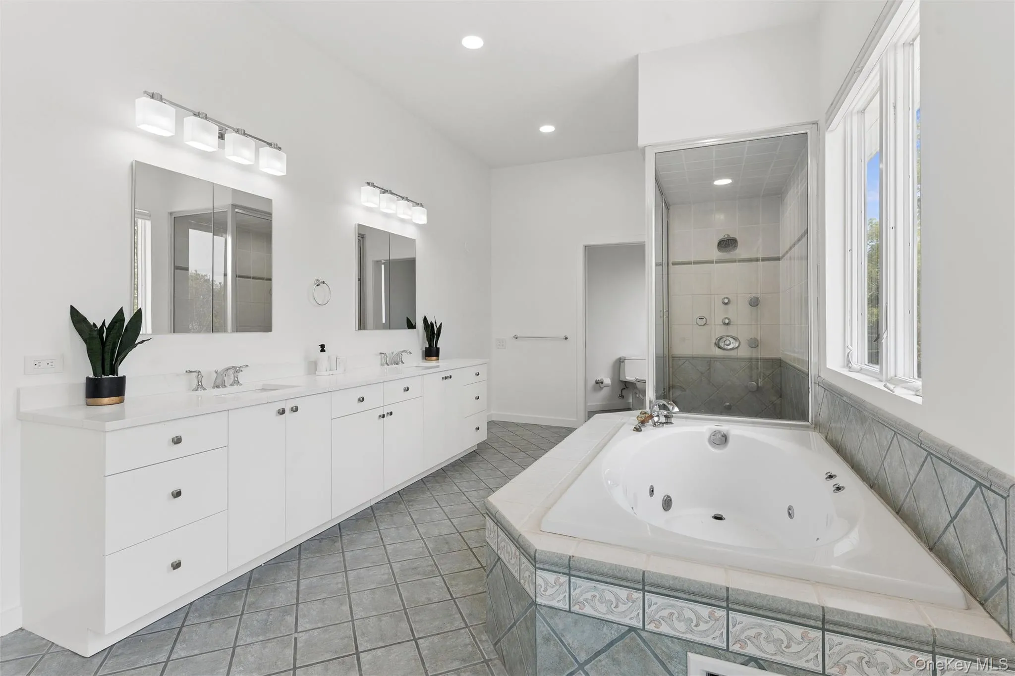 Spacious bathroom featuring a built-in jetted tub, glass-enclosed shower, dual vanities with white cabinetry, and large windows Spacious bathroom featuring a built-in jetted tub, glass-enclosed shower, dual vanities with white cabinetry, and large windows