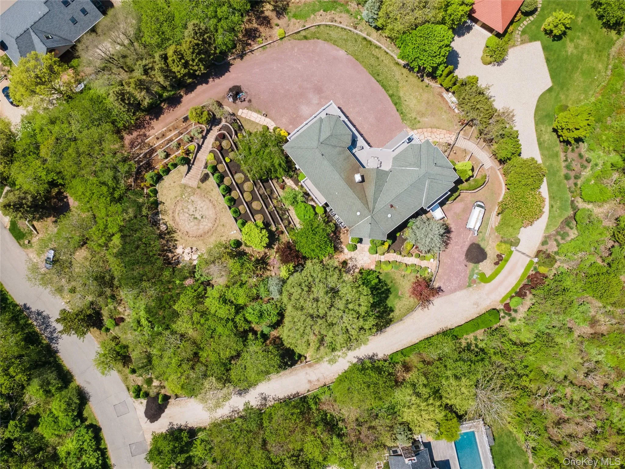 Expansive property featuring a multi-level home with a green shingled roof, a paved circular driveway, mature trees, and tiered landscaping Expansive property featuring a multi-level home with a green shingled roof, a paved circular driveway, mature trees, and tiered landscaping