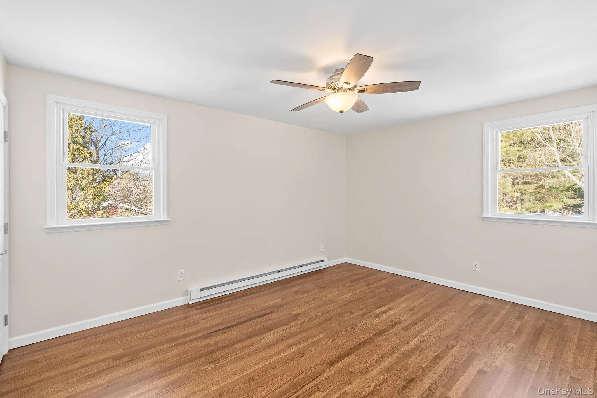 Unfurnished room featuring hardwood / wood-style flooring, a baseboard radiator, a healthy amount of sunlight, and ceiling fan Unfurnished room featuring hardwood / wood-style flooring, a baseboard radiator, a healthy amount of sunlight, and ceiling fan