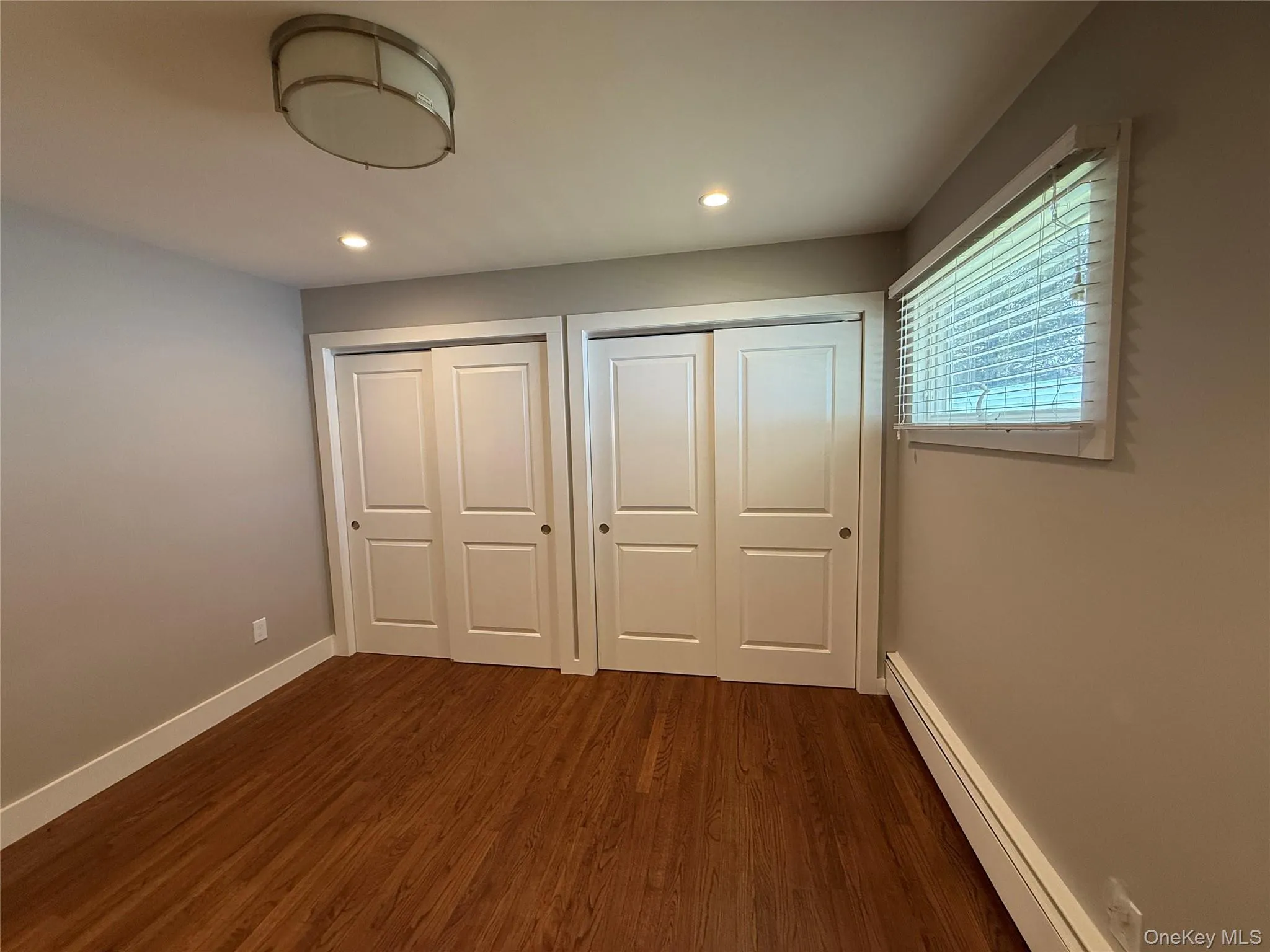 Unfurnished bedroom with baseboards, two closets, baseboard heating, and dark wood-style floors Unfurnished bedroom with baseboards, two closets, baseboard heating, and dark wood-style floors