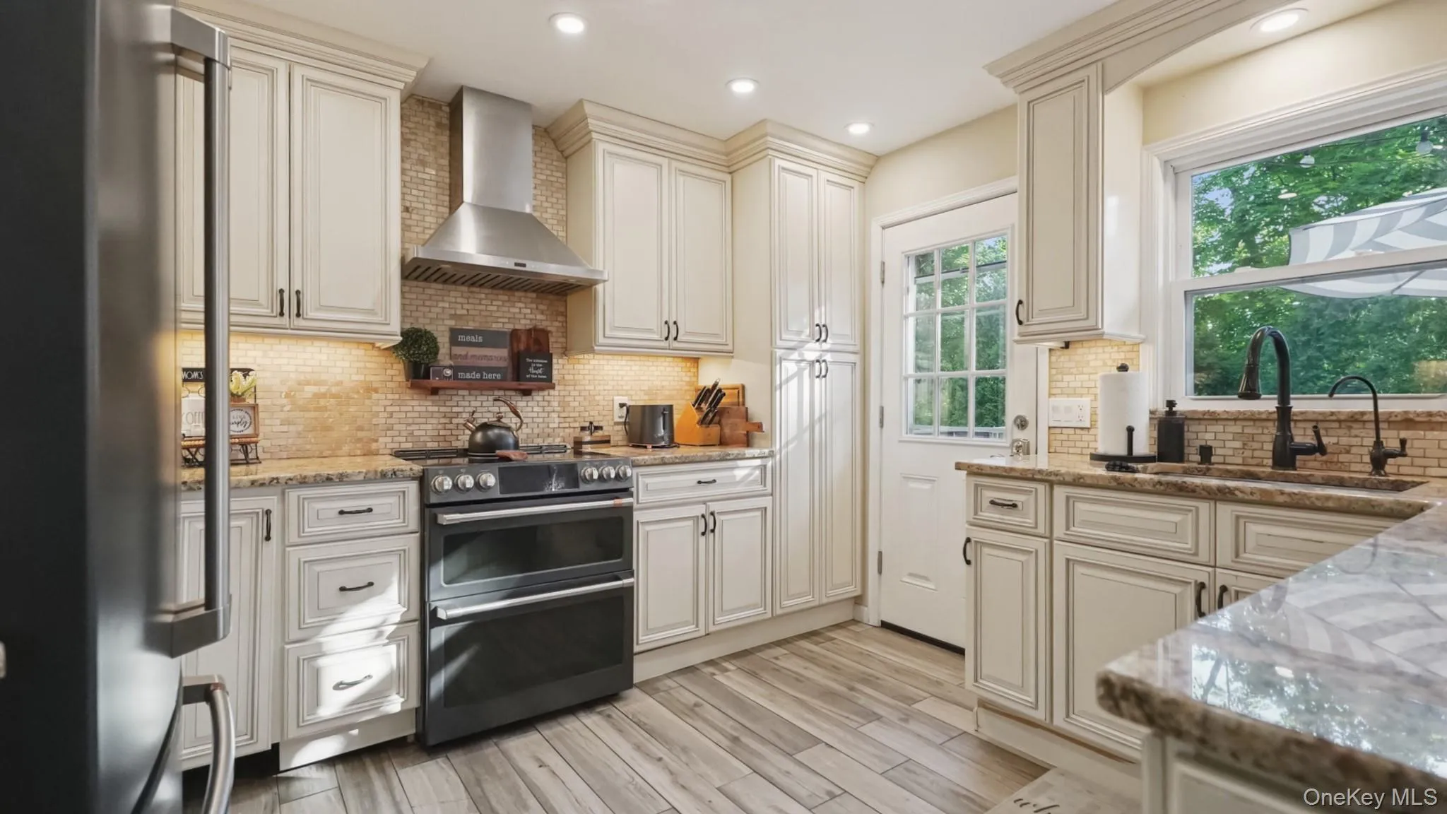 Kitchen featuring light-toned cabinetry, wood-finish flooring, a black-finish refrigerator, and a built-in range with an overhead exhaust fan Kitchen featuring light-toned cabinetry, wood-finish flooring, a black-finish refrigerator, and a built-in range with an overhead exhaust fan