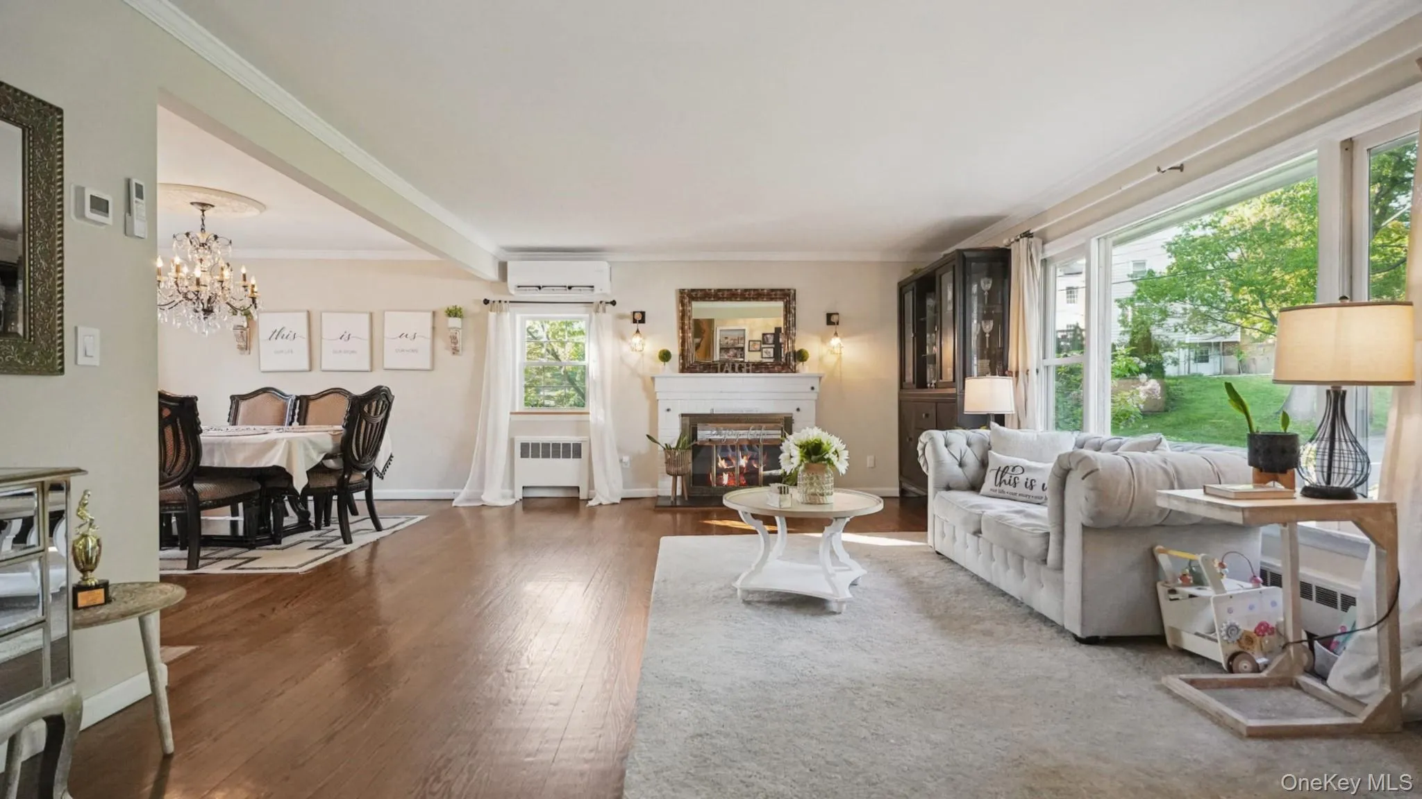 Bright living area featuring a fireplace with a decorative mantel, wood-finish flooring, a wall-mounted air conditioning unit, and large windows Bright living area featuring a fireplace with a decorative mantel, wood-finish flooring, a wall-mounted air conditioning unit, and large windows