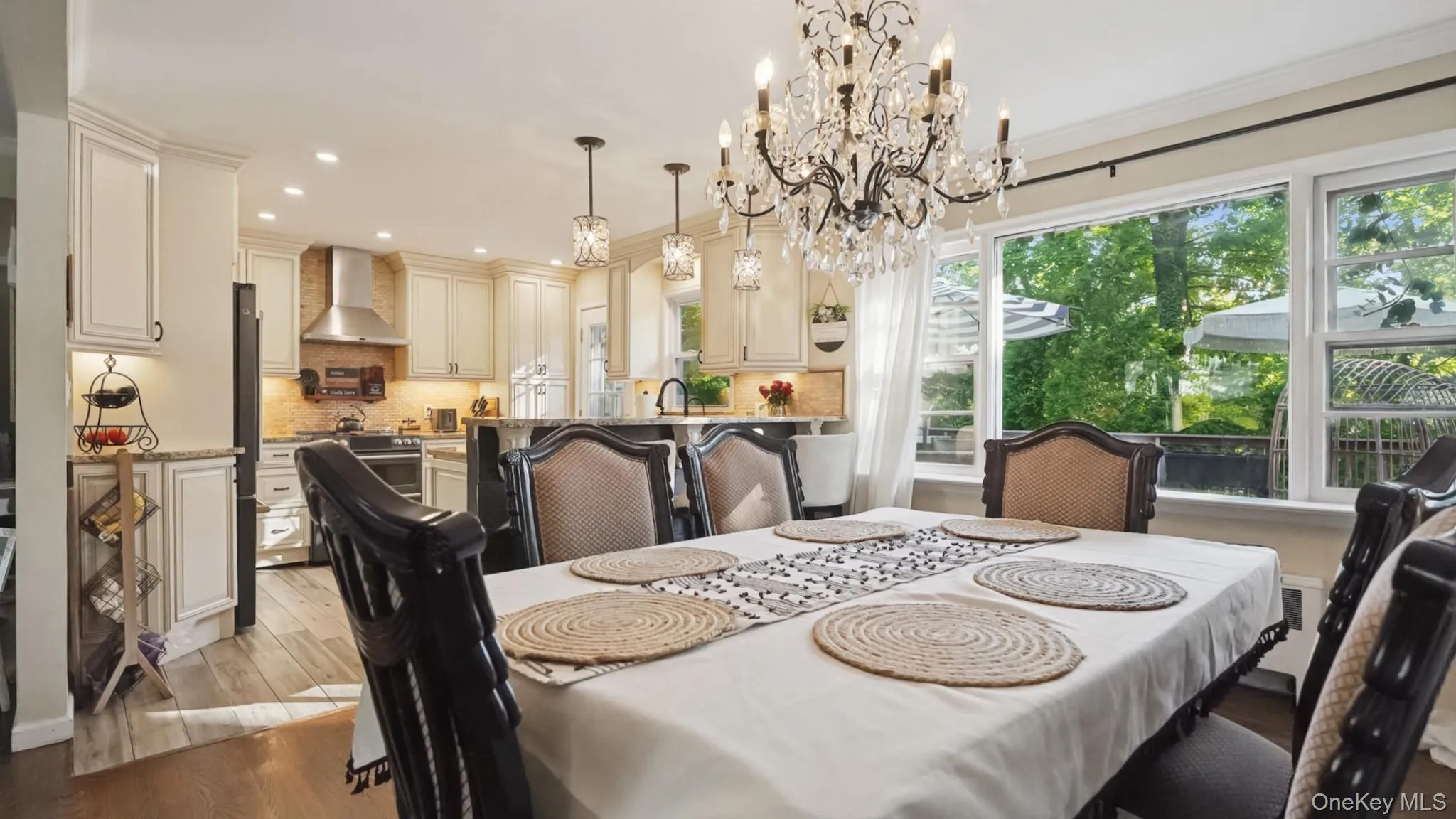 Open-concept dining area with a crystal chandelier, hardwood flooring, and a kitchen featuring light-toned cabinetry, stainless steel appliances, and a tiled backsplash Open-concept dining area with a crystal chandelier, hardwood flooring, and a kitchen featuring light-toned cabinetry, stainless steel appliances, and a tiled backsplash