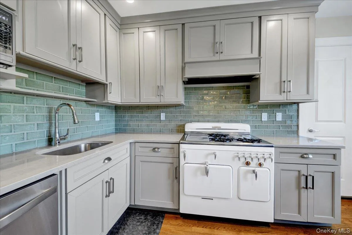 Kitchen featuring light gray shaker-style cabinetry with varied hardware, light-toned countertops, and a blue-green brick-patterned backsplash Kitchen featuring light gray shaker-style cabinetry with varied hardware, light-toned countertops, and a blue-green brick-patterned backsplash