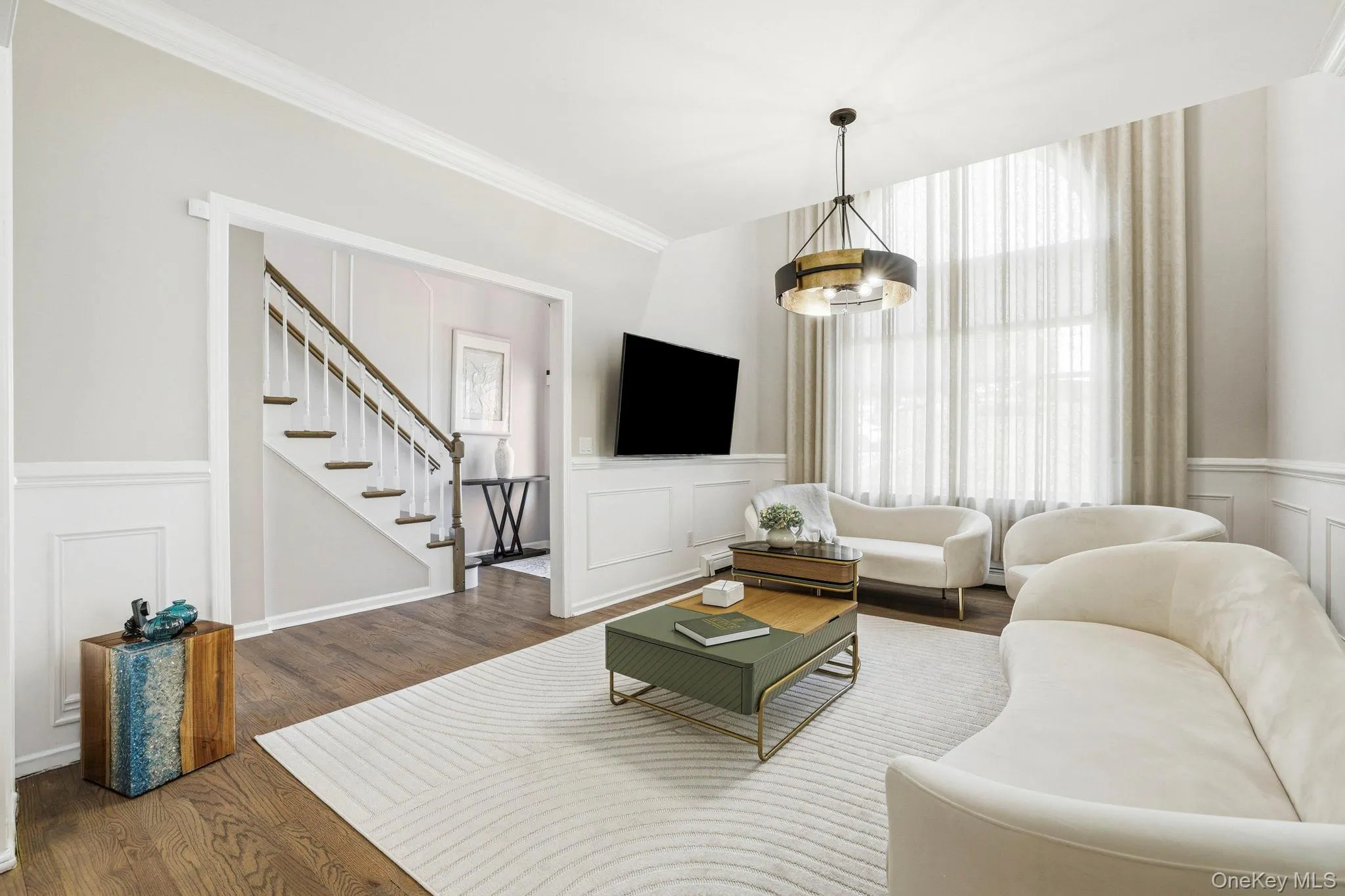 Living area featuring wood-finish flooring, wainscoting, crown molding, a decorative chandelier, and large windows with sheer and opaque drapery Living area featuring wood-finish flooring, wainscoting, crown molding, a decorative chandelier, and large windows with sheer and opaque drapery