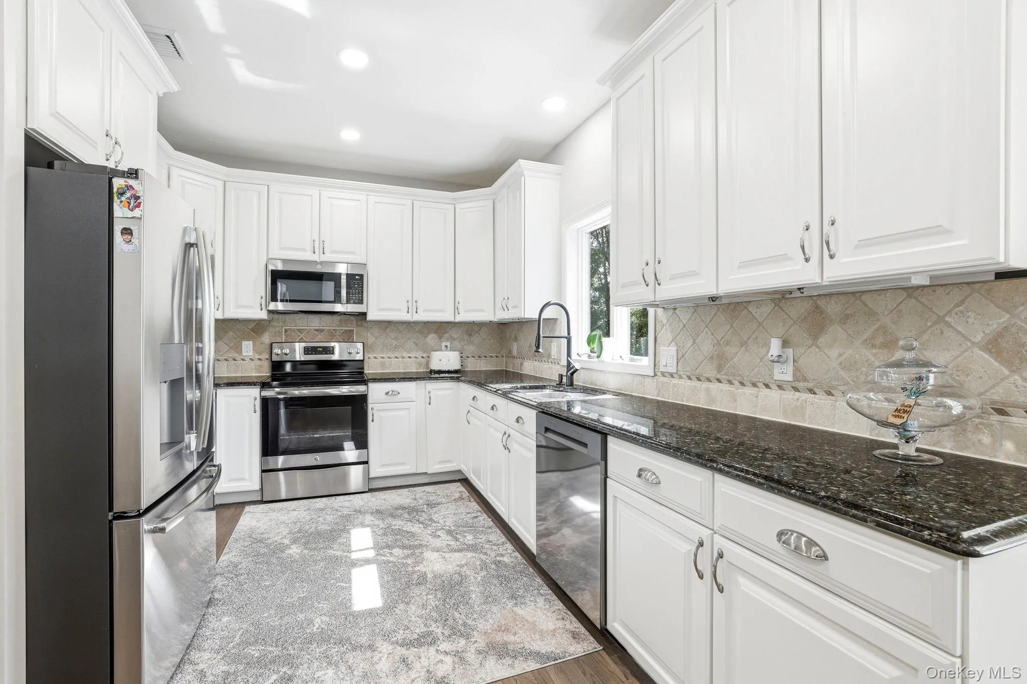Bright kitchen featuring white cabinetry, dark stone countertops, stainless steel appliances, and a tiled backsplash Bright kitchen featuring white cabinetry, dark stone countertops, stainless steel appliances, and a tiled backsplash