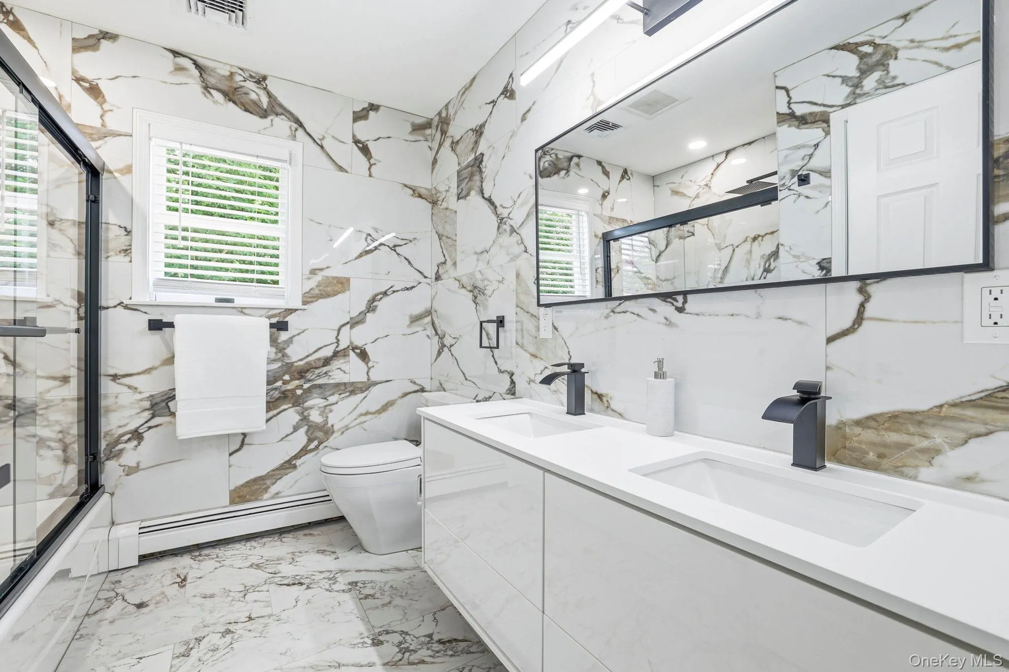 Bathroom featuring full-height marble-look tile, a dual-sink vanity with integrated white countertops, and matte black fixtures Bathroom featuring full-height marble-look tile, a dual-sink vanity with integrated white countertops, and matte black fixtures