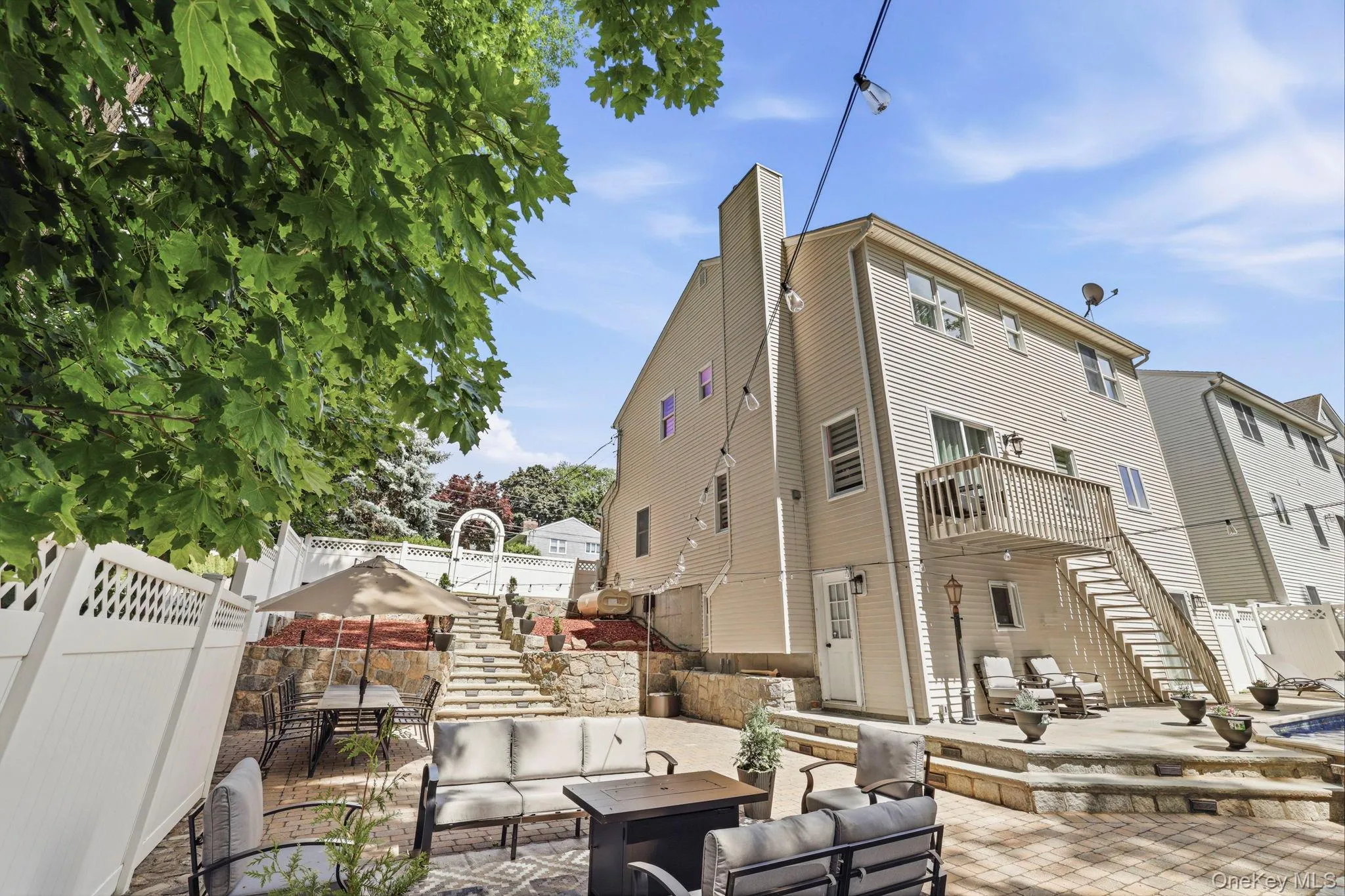 Multi-level outdoor entertaining area featuring a paver patio with stone retaining walls, a deck with stairs to the yard, white lattice fencing, and an arched garden trellis Multi-level outdoor entertaining area featuring a paver patio with stone retaining walls, a deck with stairs to the yard, white lattice fencing, and an arched garden trellis