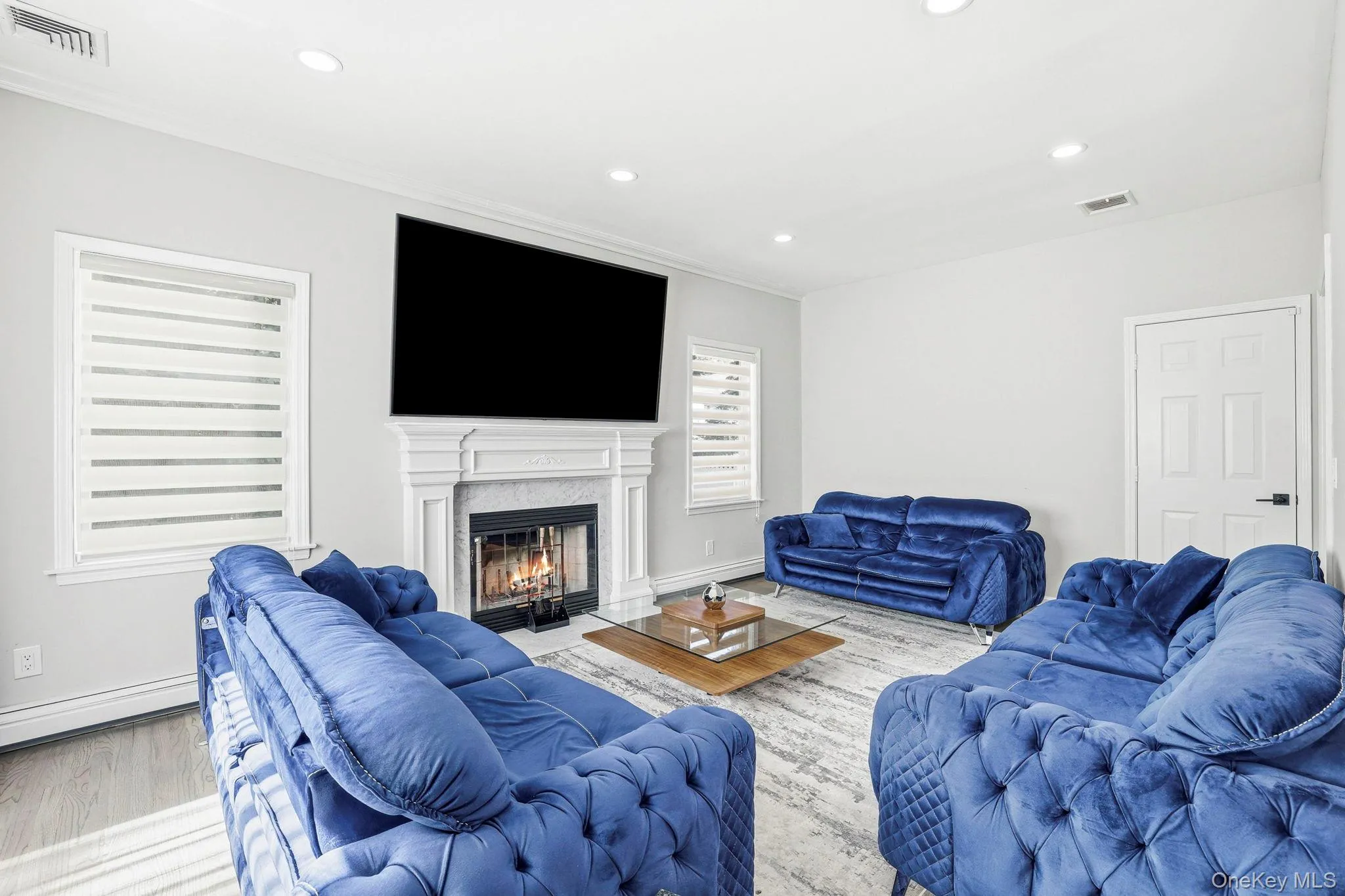 Living space featuring a detailed fireplace surround, recessed lighting, wood-finish flooring, and crown molding Living space featuring a detailed fireplace surround, recessed lighting, wood-finish flooring, and crown molding
