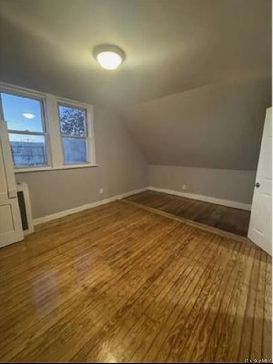 Room featuring wood-finish flooring, a sloped ceiling, two windows with white trim, a flush mount ceiling light fixture, and white baseboards Room featuring wood-finish flooring, a sloped ceiling, two windows with white trim, a flush mount ceiling light fixture, and white baseboards