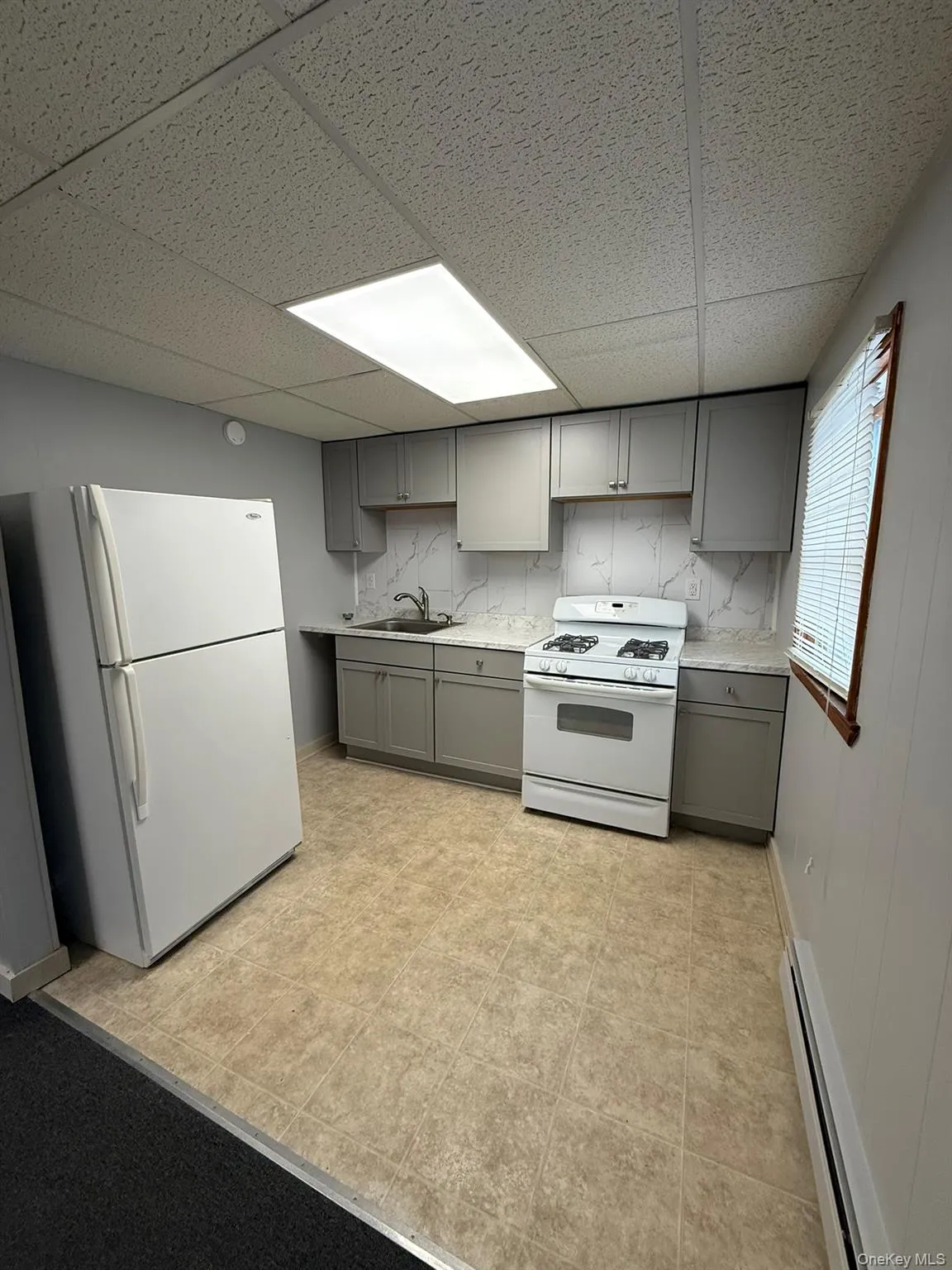 Kitchen featuring neutral-toned cabinetry, light-toned countertops, tile-patterned flooring, a recessed ceiling light fixture, and a window with blinds Kitchen featuring neutral-toned cabinetry, light-toned countertops, tile-patterned flooring, a recessed ceiling light fixture, and a window with blinds